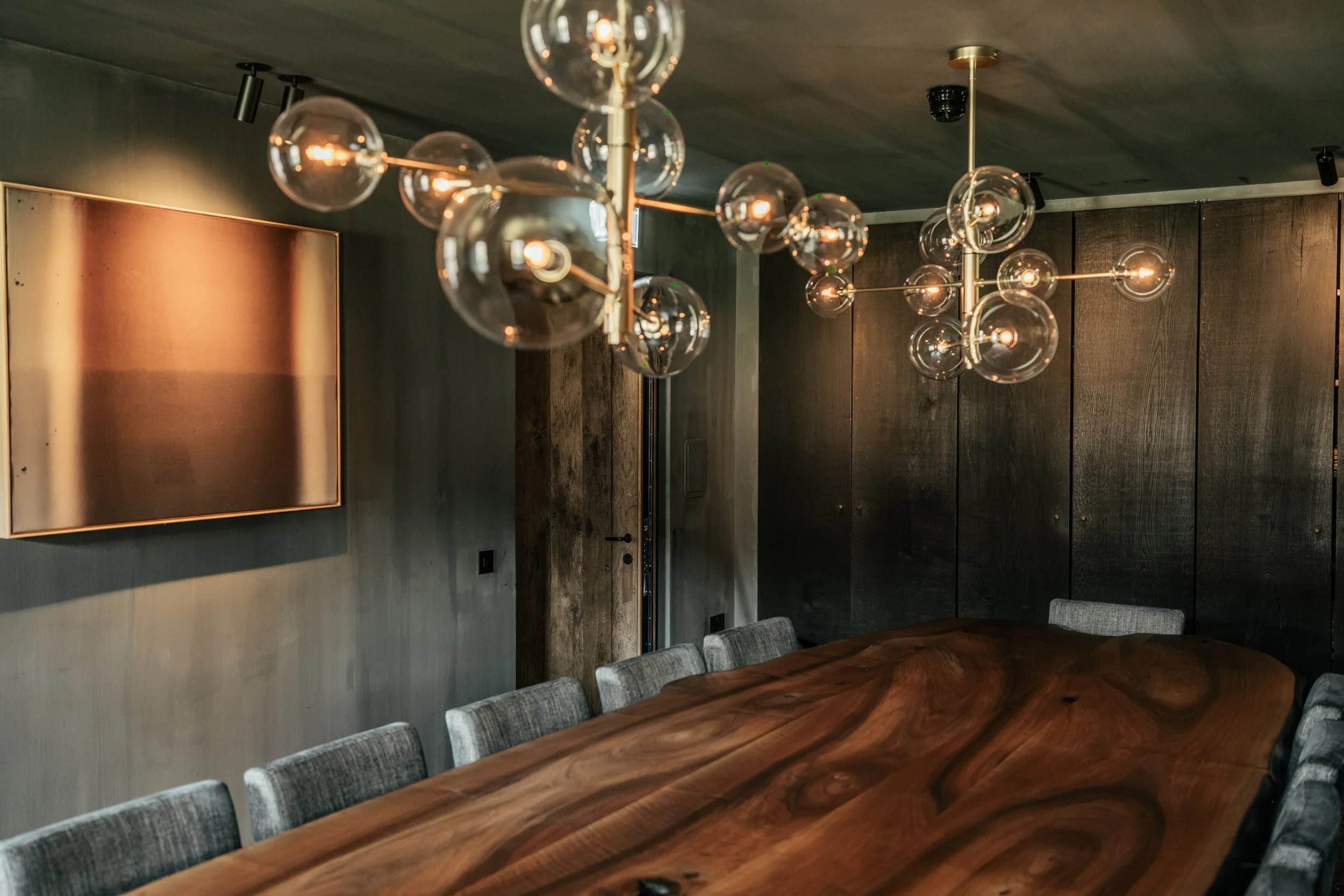 Interior of a modern dining room with a large wooden table surrounded by gray upholstered chairs, dark wooden wall panels, and a chandelier with multiple spherical glass bulbs hanging from the ceiling.