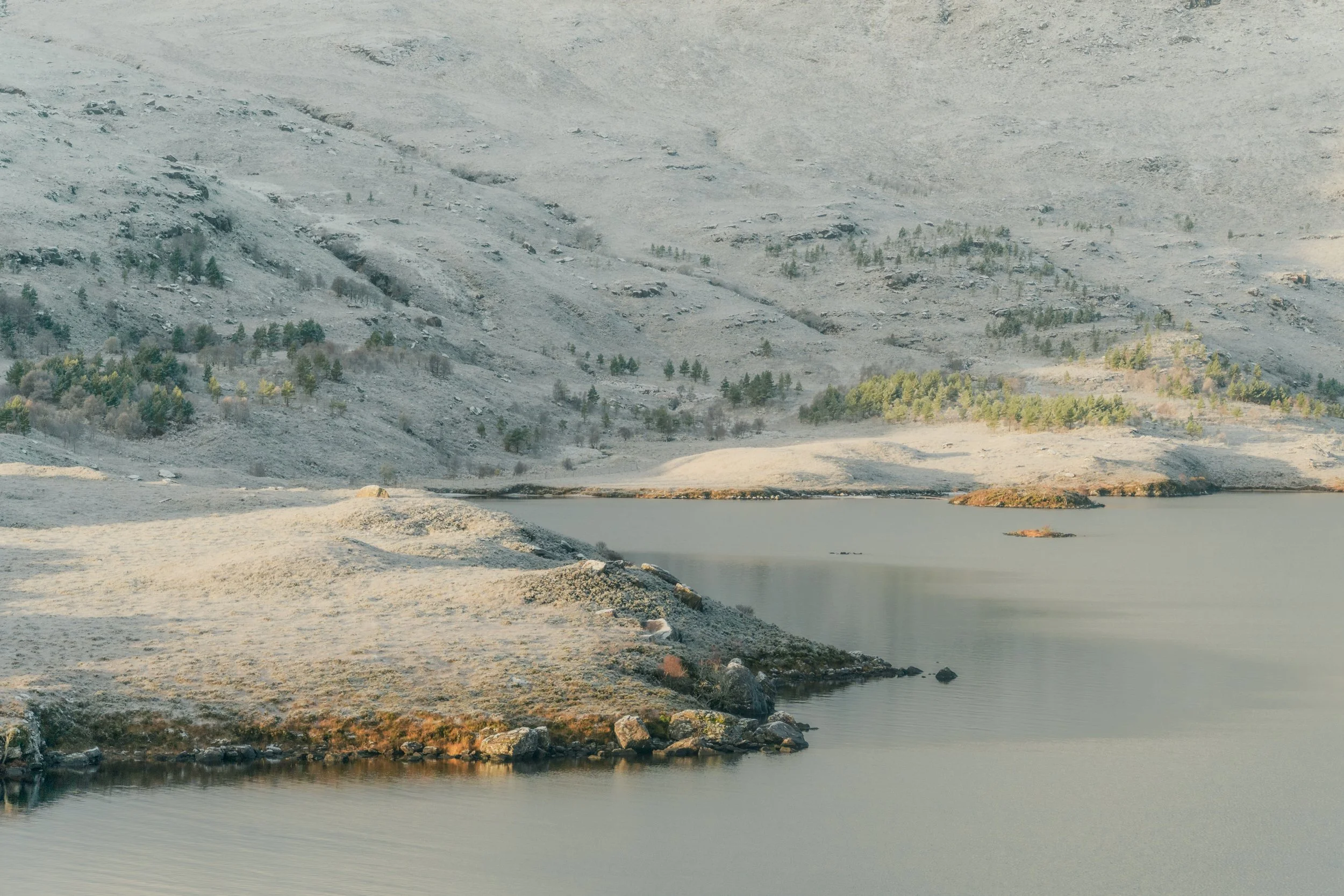 A serene landscape featuring a calm body of water in the foreground, bordered by rocky and sandy terrain, with a hillside covered in sparse trees and vegetation in the background.