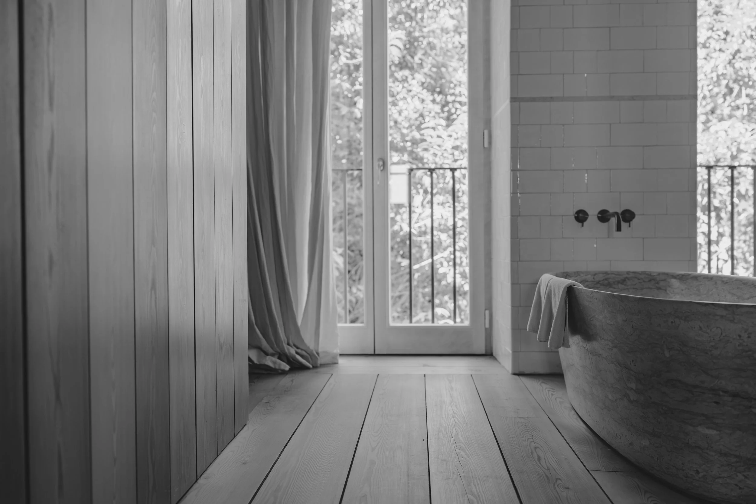 Modern bathroom with wooden floor, white brick wall, bathtub, and a large window with curtains, overlooking trees outside.