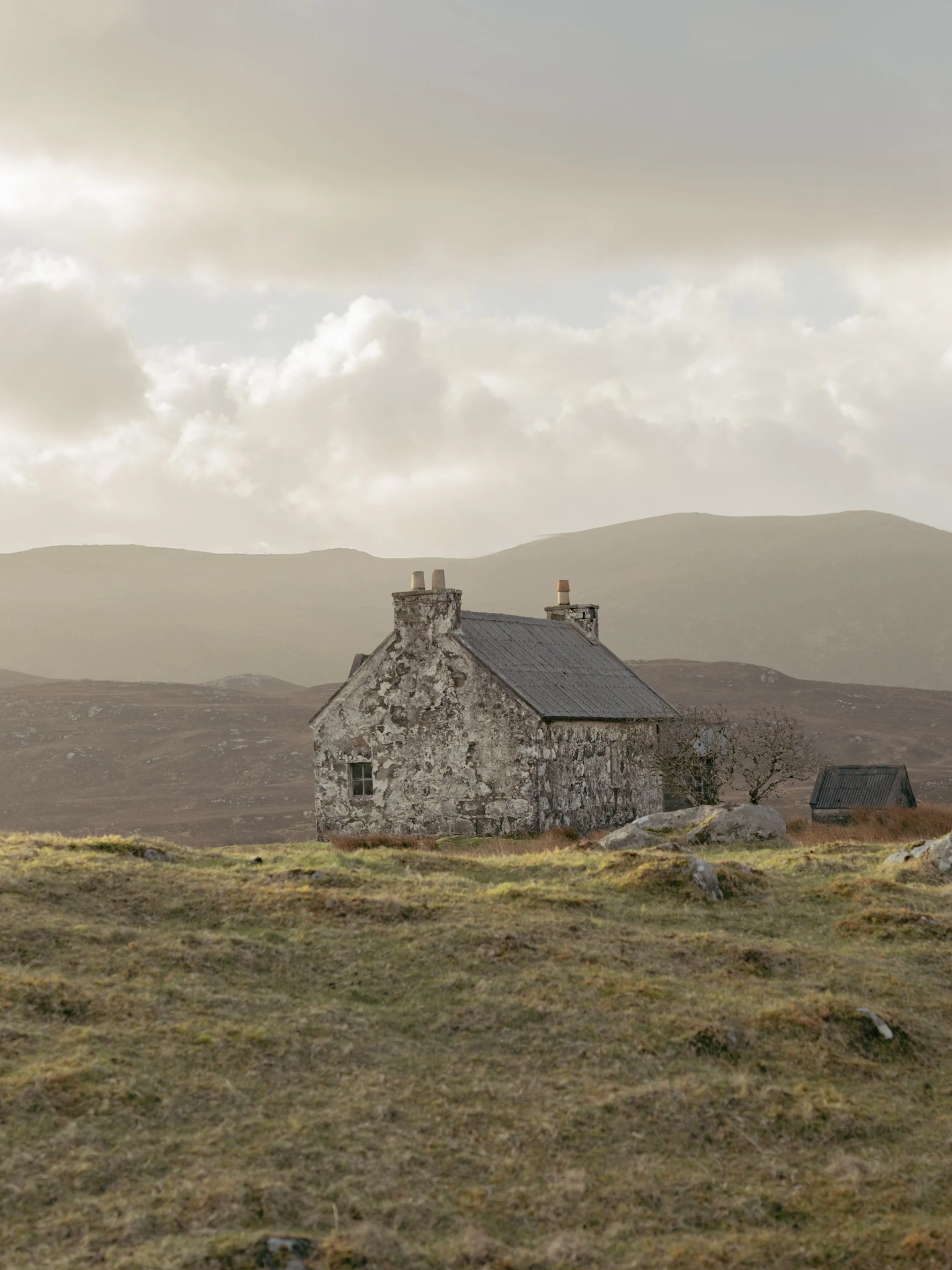 An old stone house with a gray roof in a rural landscape under a cloudy sky with distant mountains.