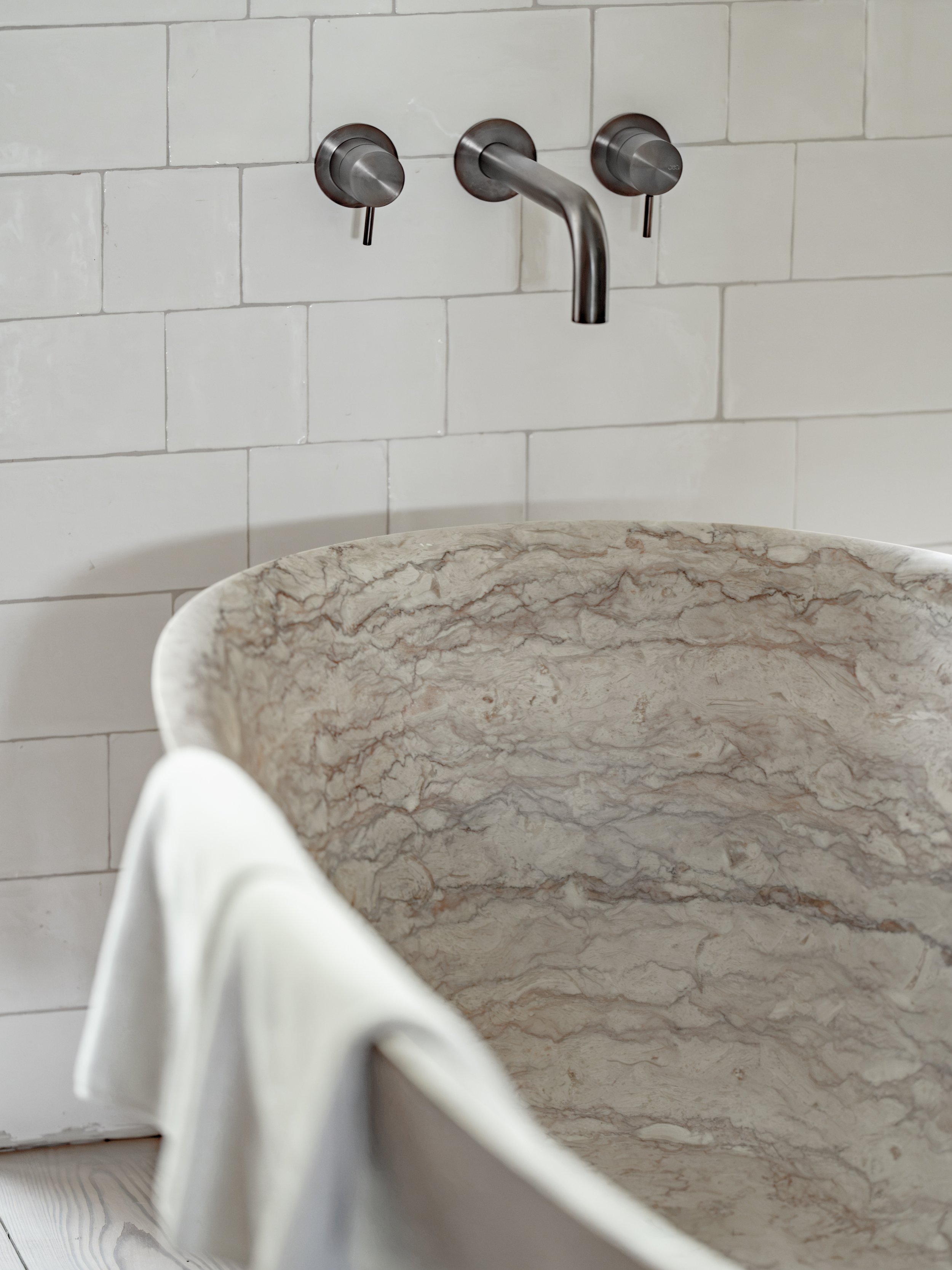 Close-up of a wall-mounted stainless steel faucet with two handles, above a marble sink, against a tiled white wall background.