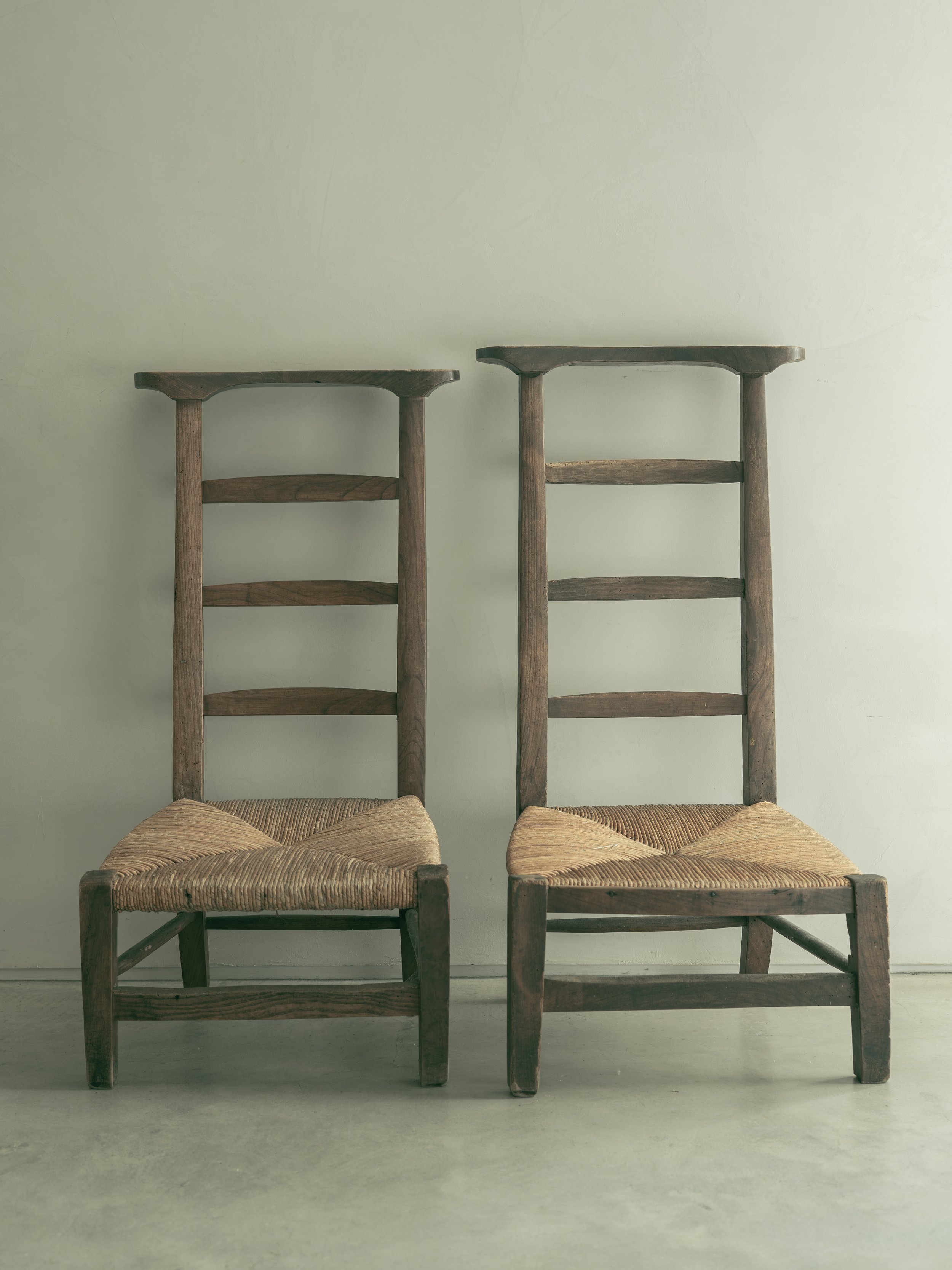 Two wooden chairs with woven seats are placed side by side against a plain, light-colored wall.