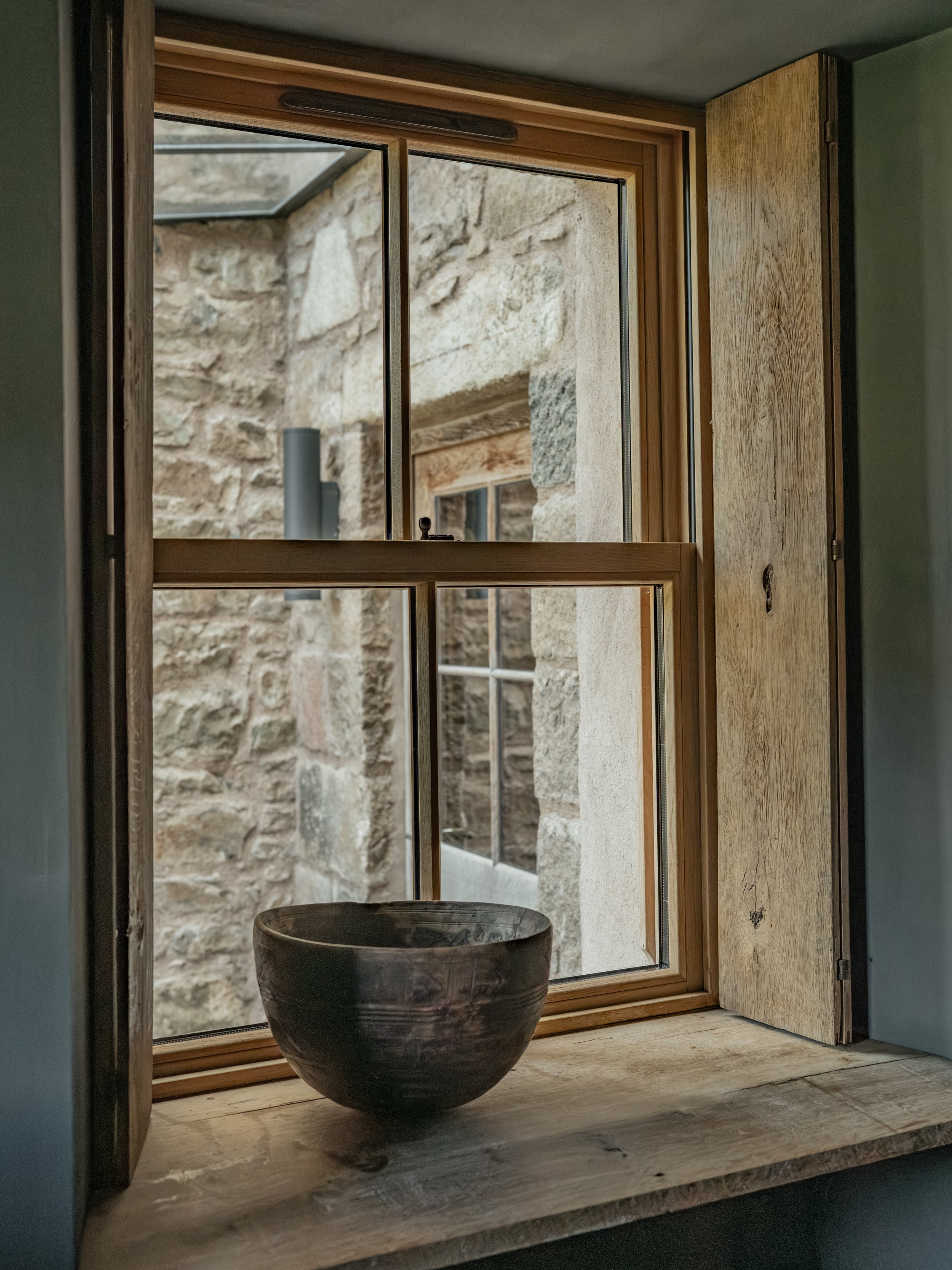 View through a window showing a stone wall outside and a pipe protruding from the wall. A wooden window frame and a wooden shutter are part of the window. A large ceramic or stone basin is on a marble or stone surface inside near the window.