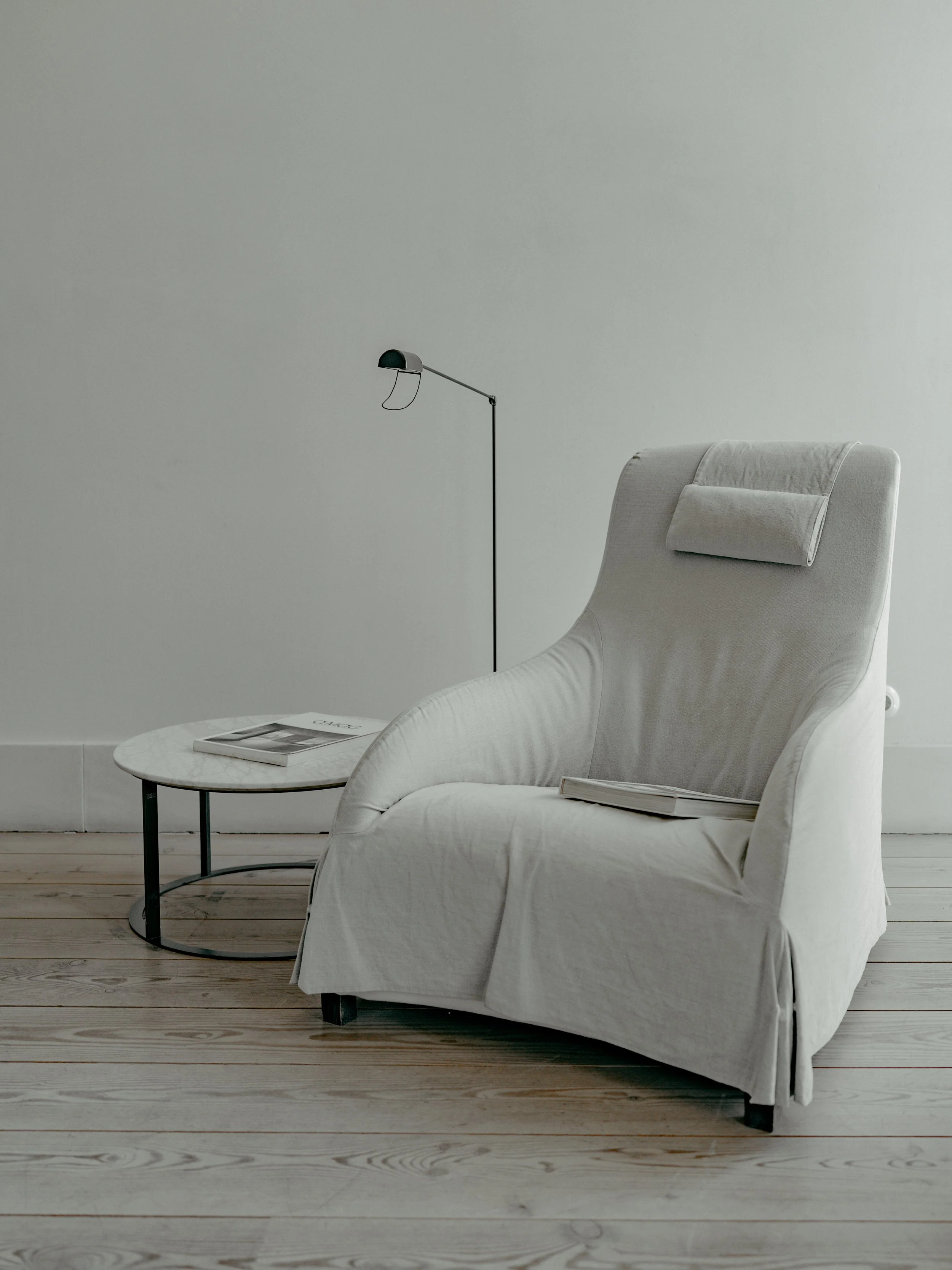 A white armchair with a pillow and a magazine on a small round black metal table in a minimalist room with gray wall and light-colored wooden floor.