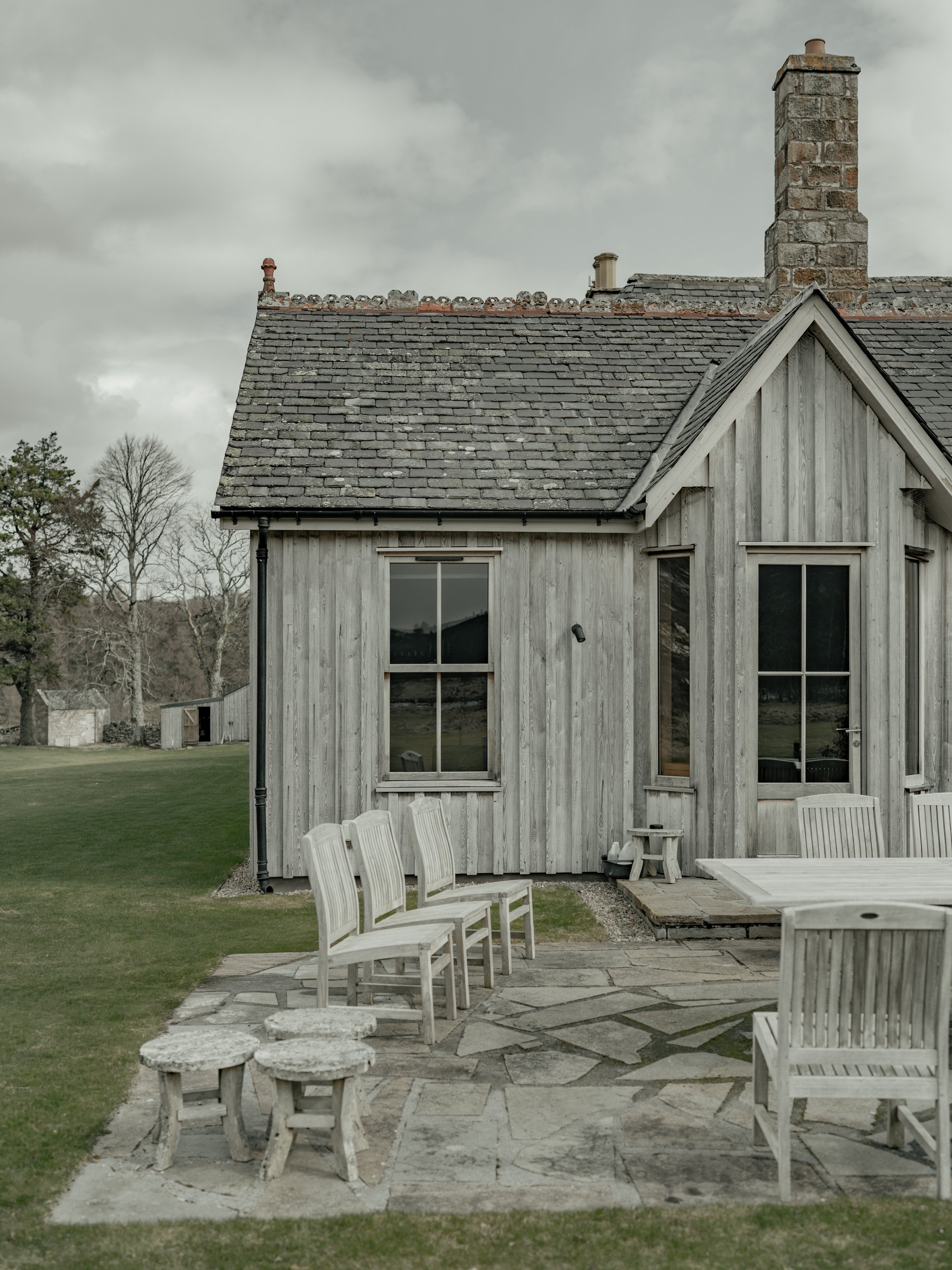 A rustic house with weathered wooden siding, a shingled roof, and brick chimneys, with outdoor furniture and a stone patio in a yard with grass and trees.