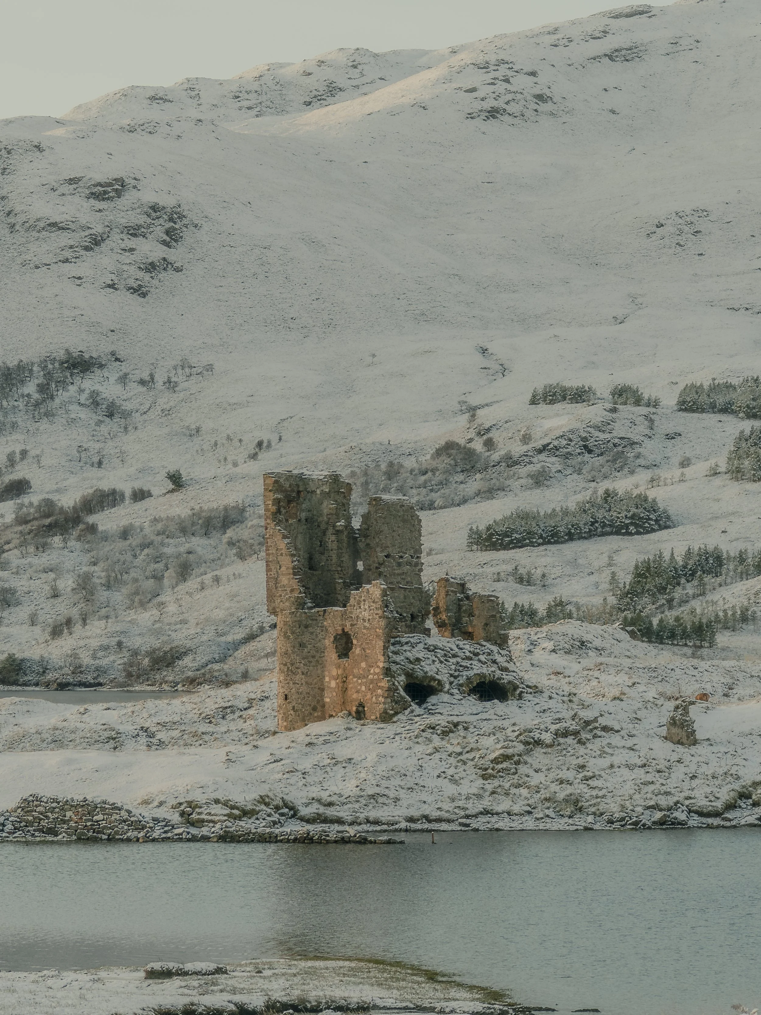 Snow-covered mountainous landscape with ruins of an old stone building near a body of water.
