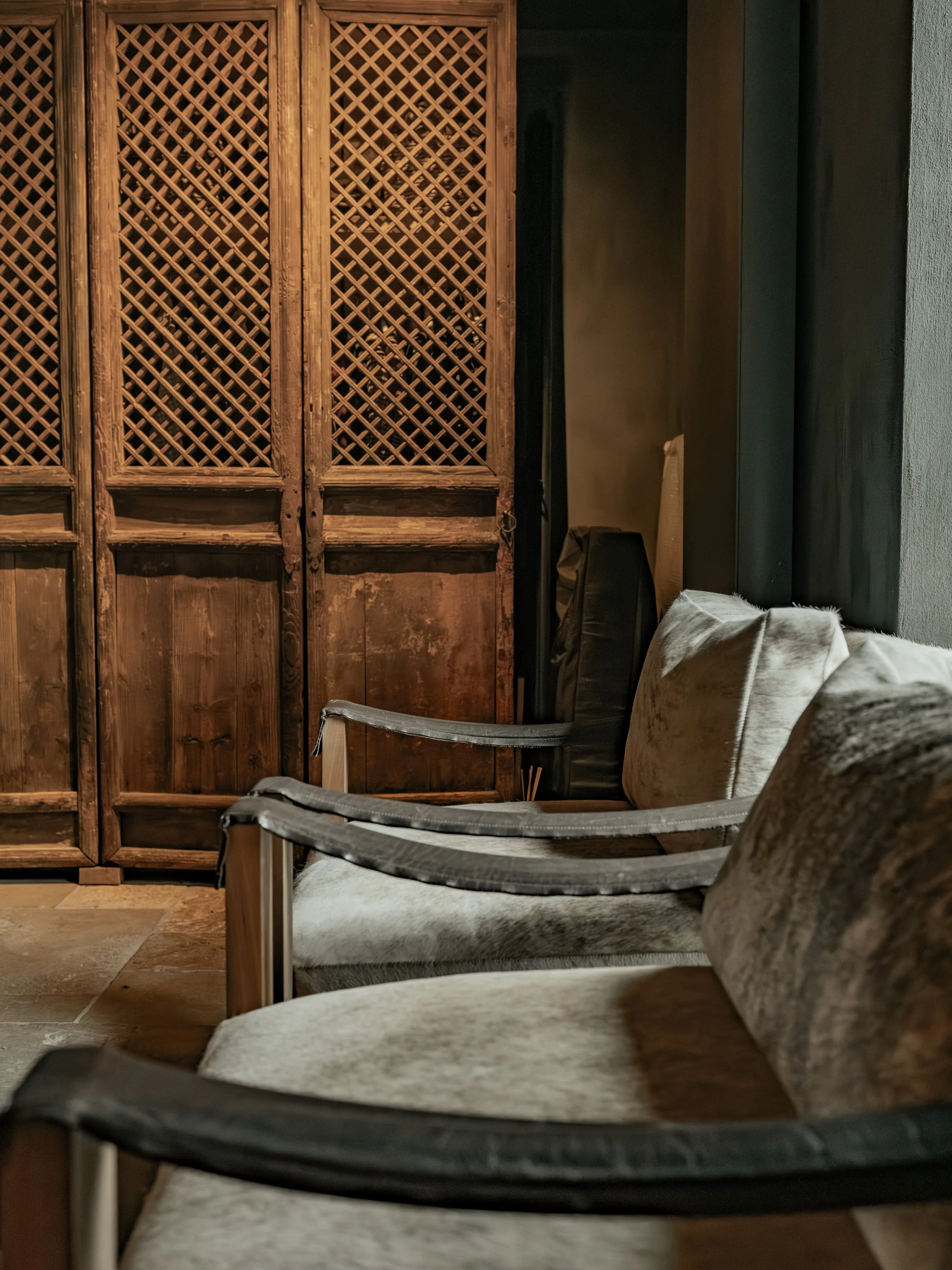 A close-up view of a rustic wooden room divider with lattice design, positioned next to a dark wall. In front of the divider are several chair-like furniture pieces with padded seats and armrests, covered in light-colored fabric.
