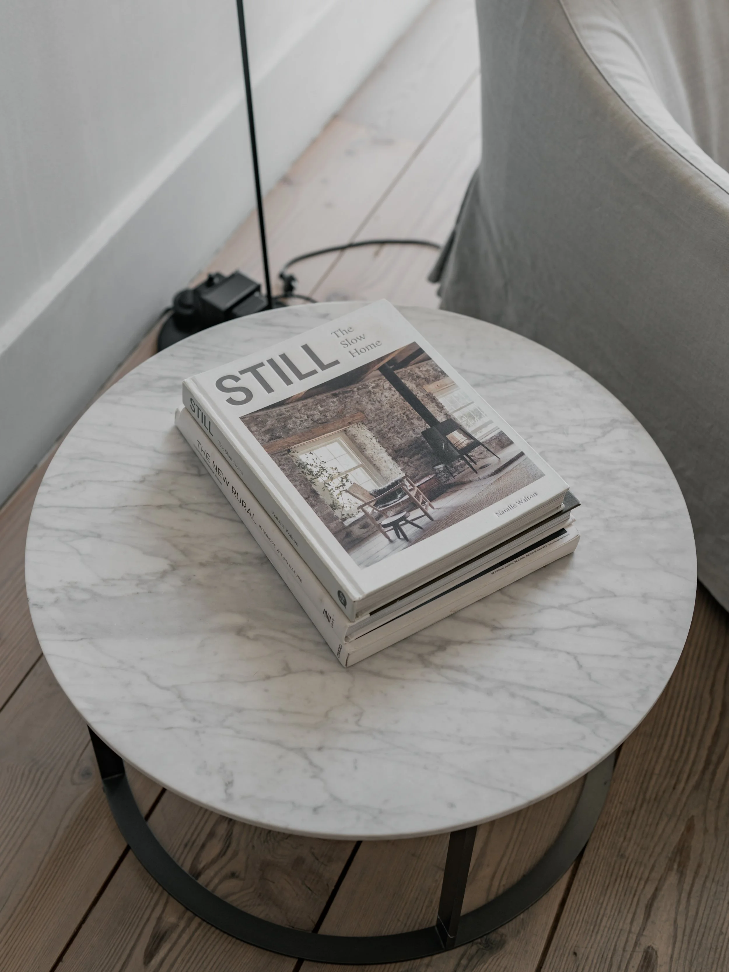 A white marble round side table with black metal legs, holding a stack of three books about rustic home design, placed next to a grey upholstered armchair on a wooden floor.