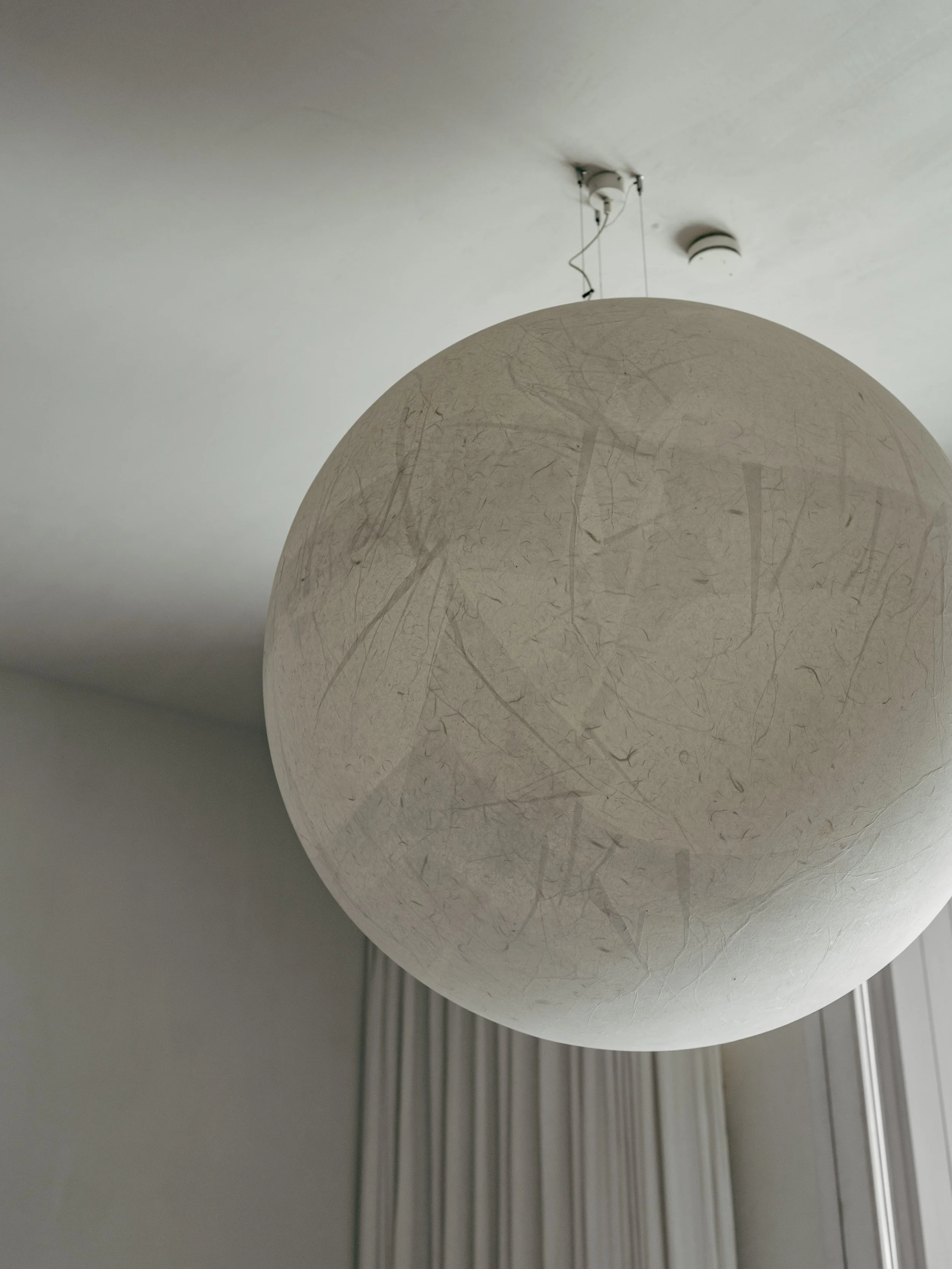 A close-up of a white spherical pendant lamp with a textured surface hanging from the ceiling. The ceiling has a smoke detector and a light fixture, with vertical blinds visible in the background.