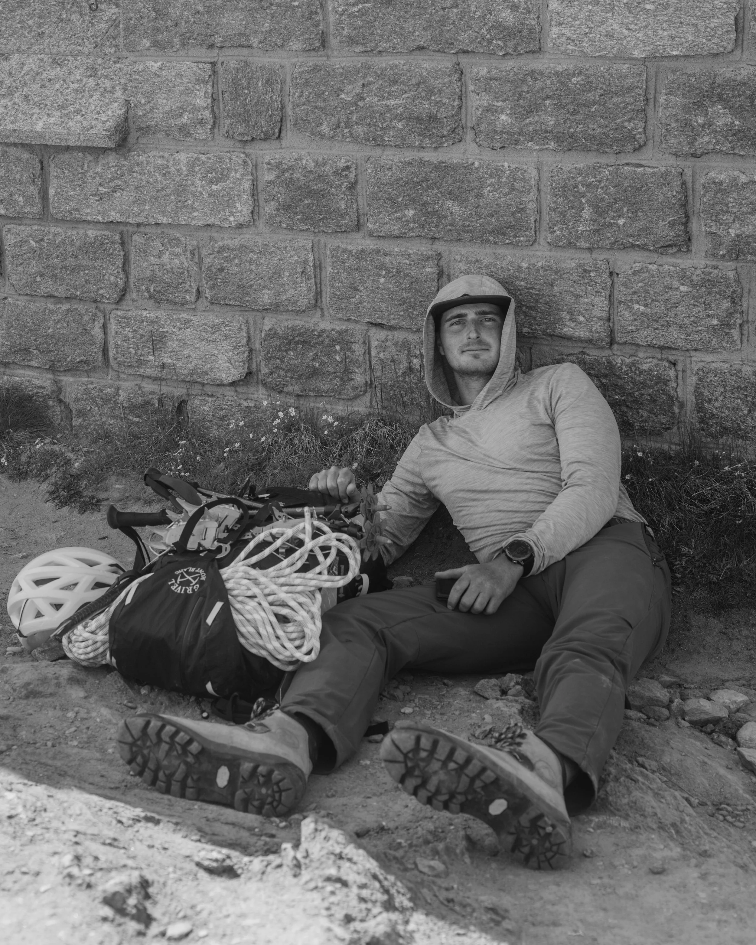 A man wearing a hoodie and outdoor gear sits leaning against a large stone brick wall, with climbing or hiking equipment, including a helmet and ropes, placed in front of him on the ground.