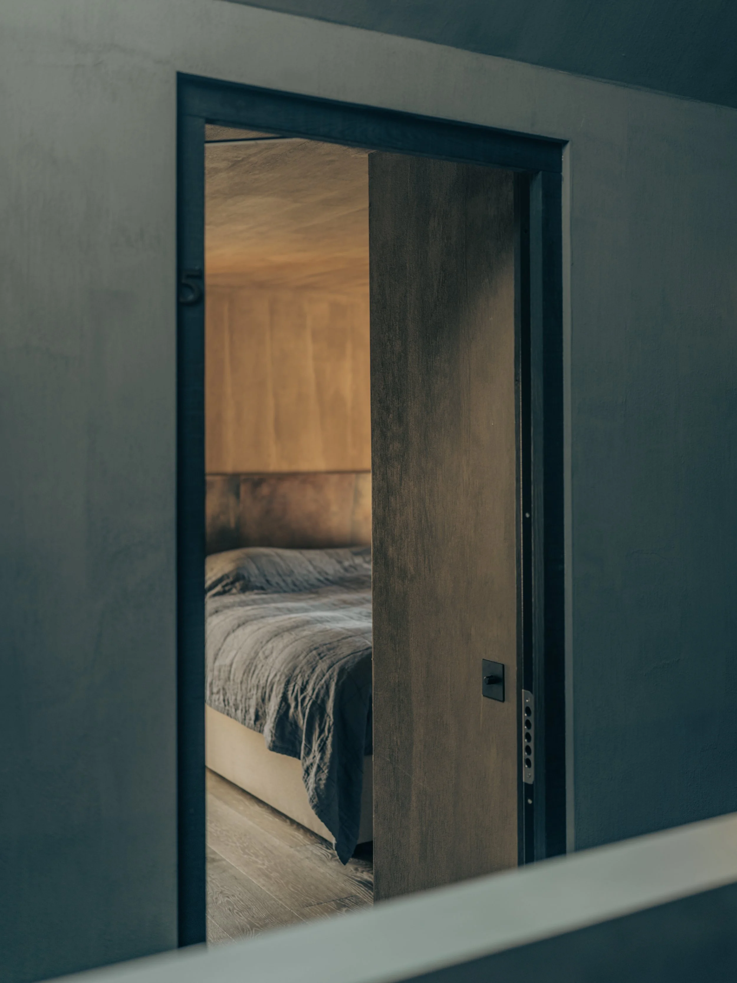 View of a bedroom seen through a slightly open door, featuring a bed with a patterned blanket and a wooden headboard.