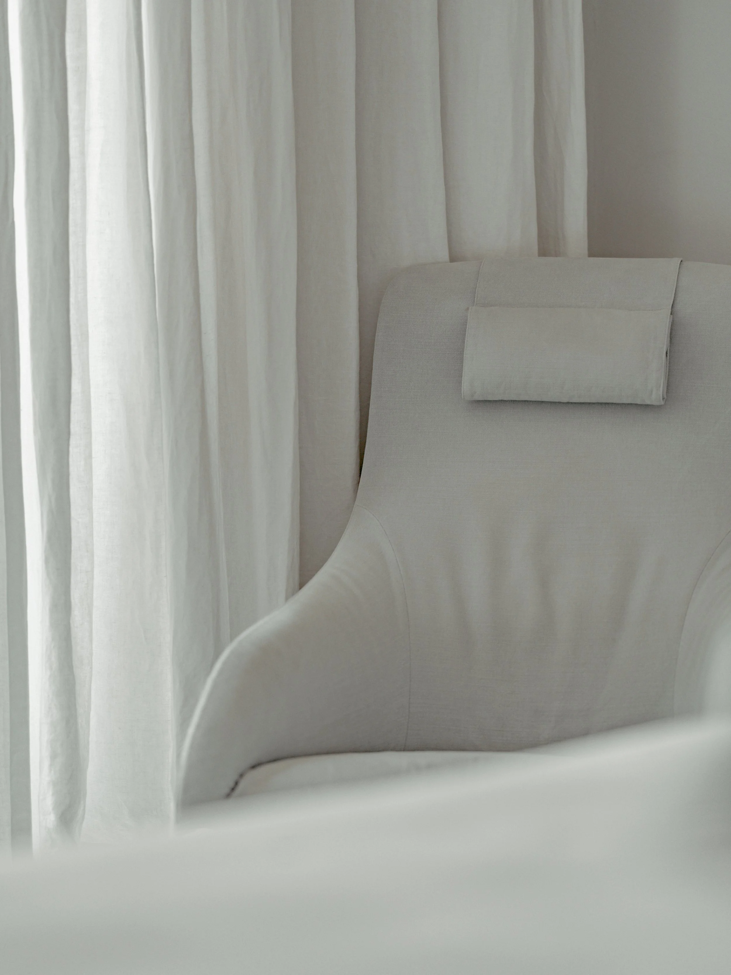 Close-up of a white curtain beside a white upholstered armchair.