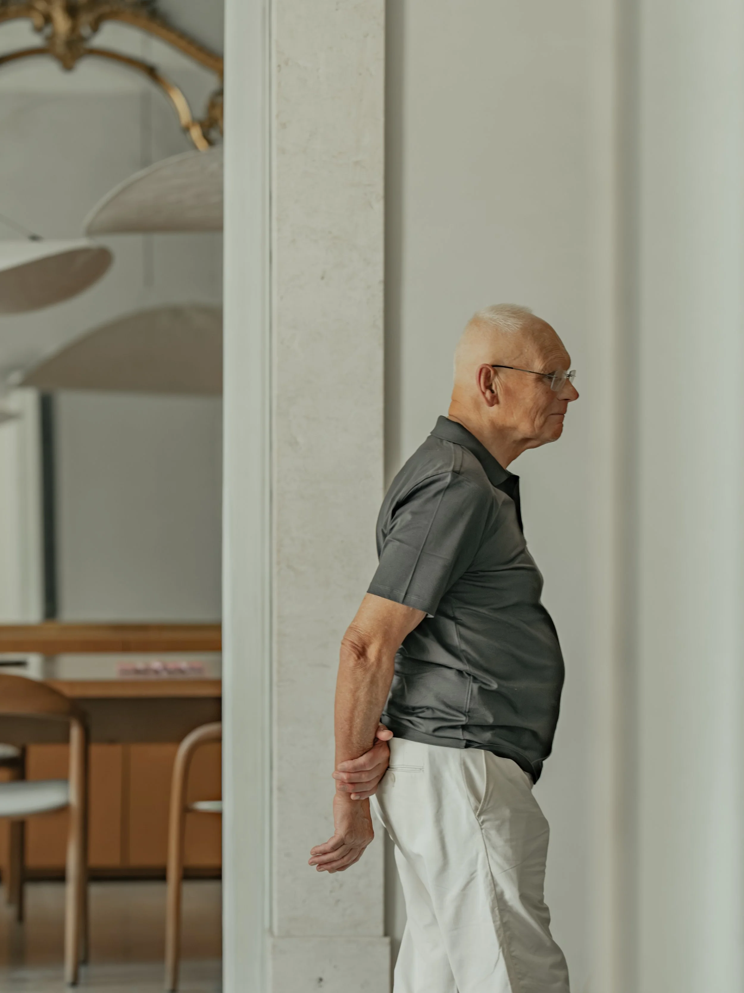 An elderly man leaning backward with his hands behind his back, standing against a wall in a room with chairs and tables in the background.