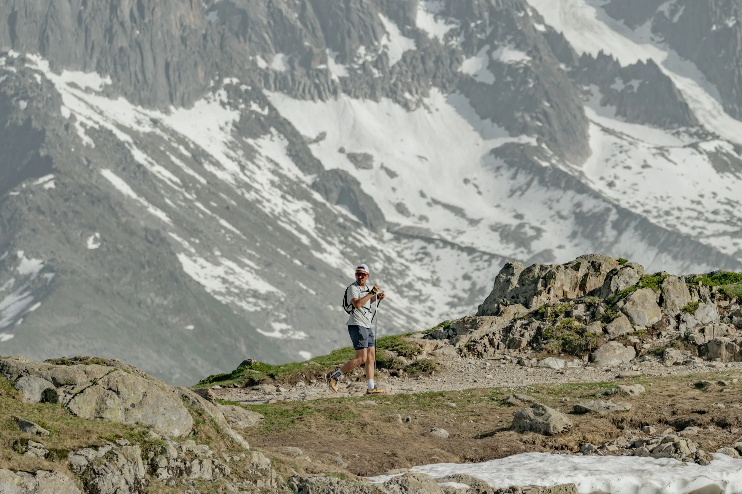 A man hiking on a rocky trail in a mountainous area with snow patches and steep rocky peaks in the background.