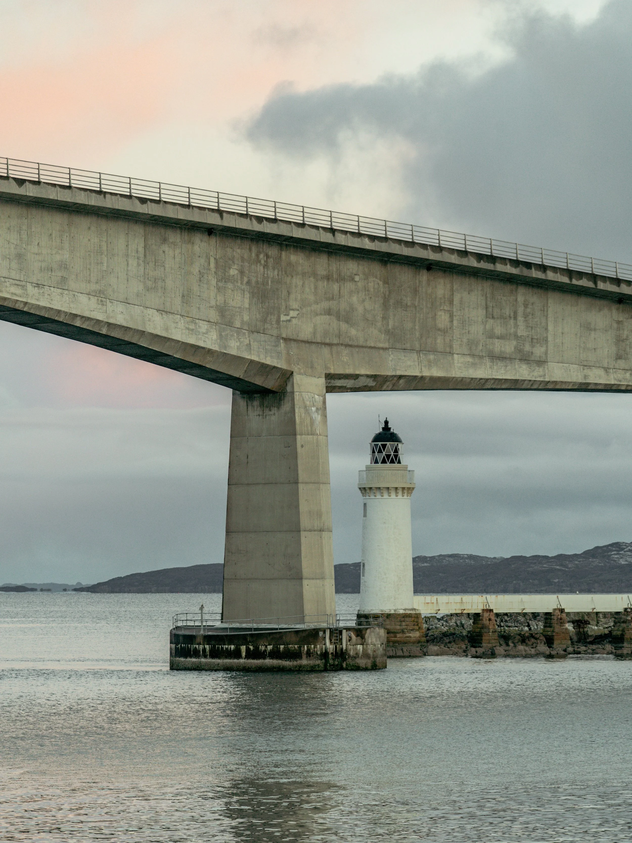 Two lighthouses standing in the water near a pier, with a concrete bridge overhead, cloudy sky, and distant land in the background.