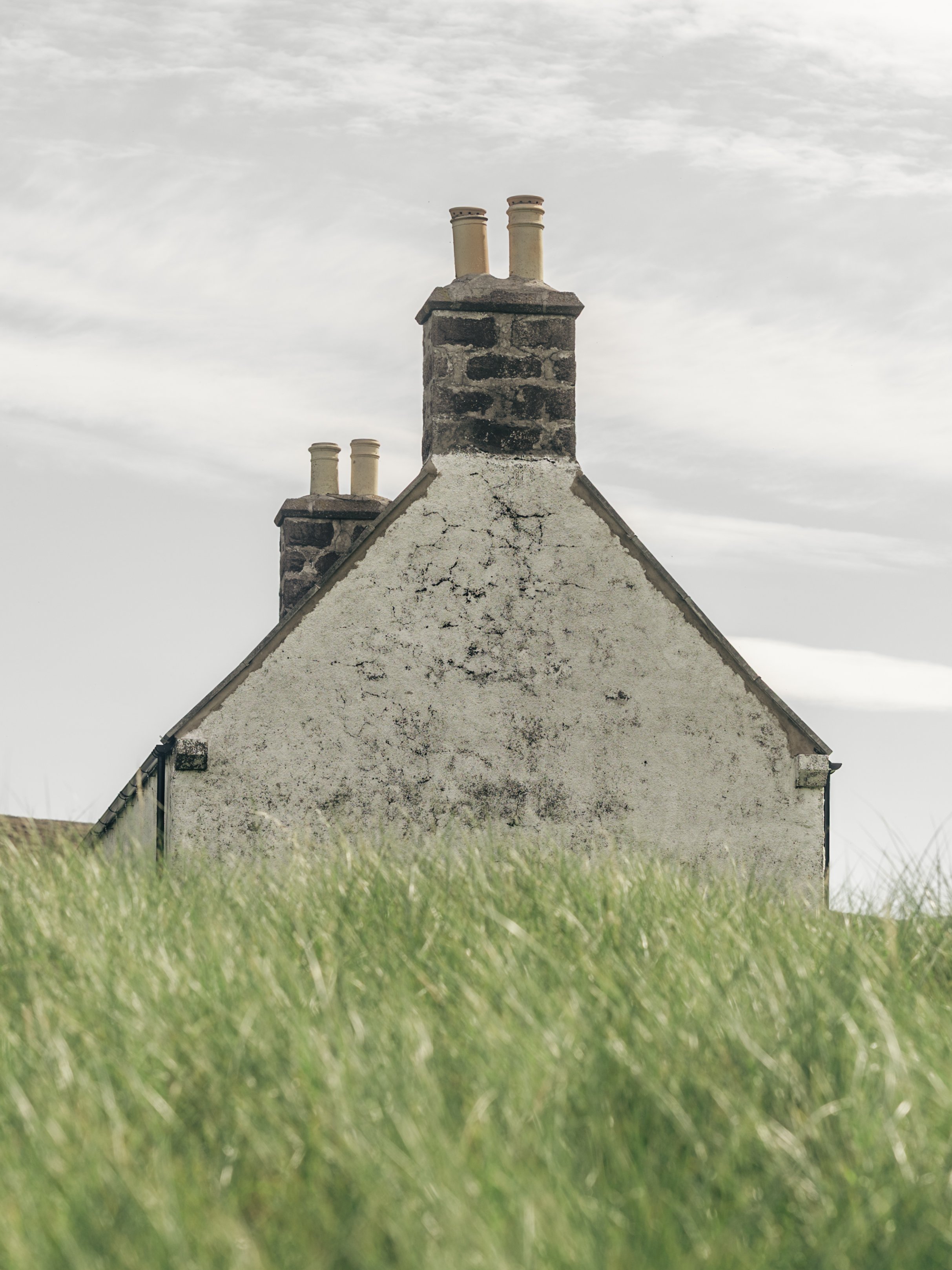 A house with a sloped roof, a prominent chimney with two stacks, and a textured exterior wall, set against a cloudy sky with grass in the foreground.