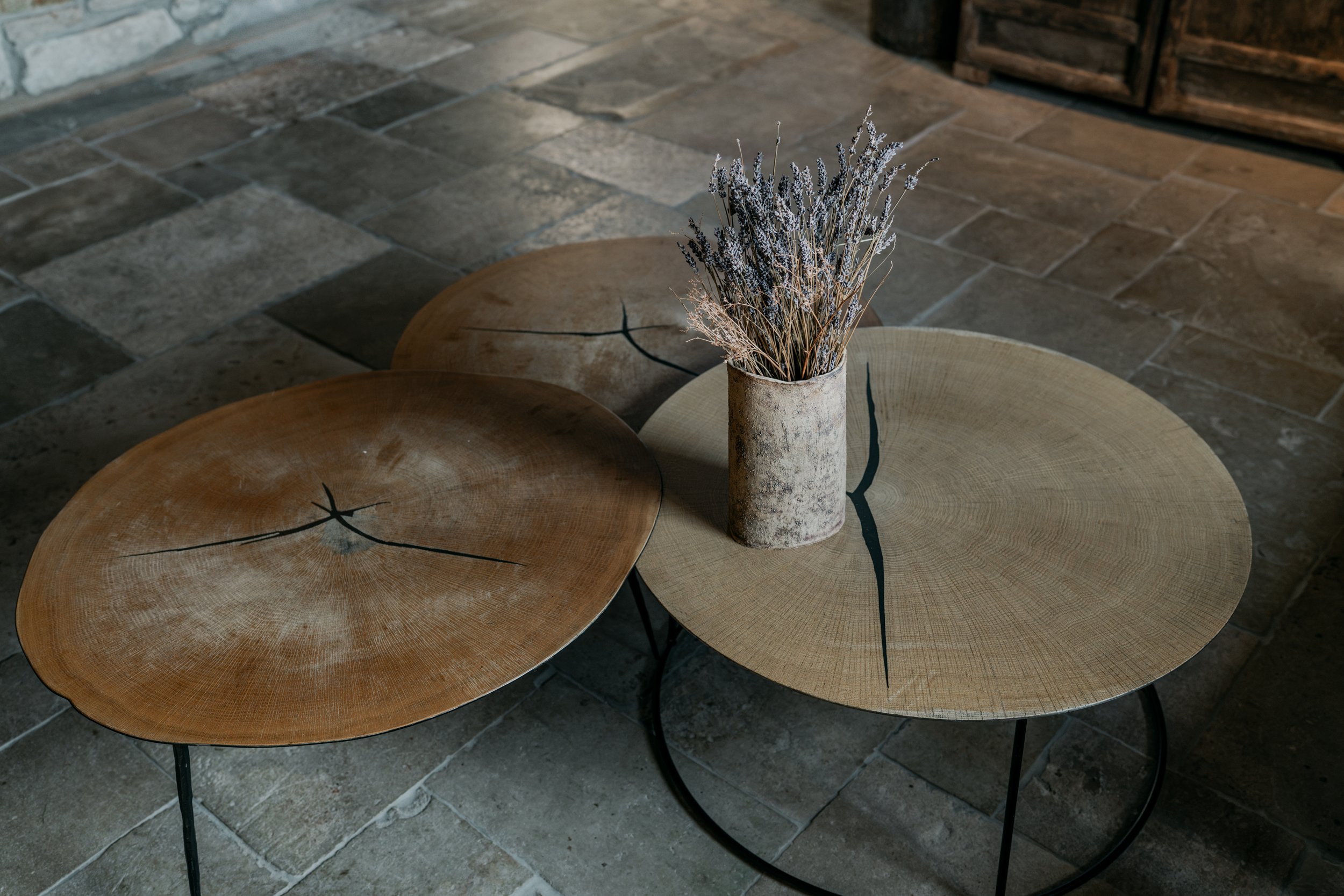 Three wooden tables with black metal legs, two with a light wood finish and one with a darker finish, arranged close together on a stone floor. A tall, cylindrical, rustic concrete vase with dried lavender flowers sits on the light finished table.