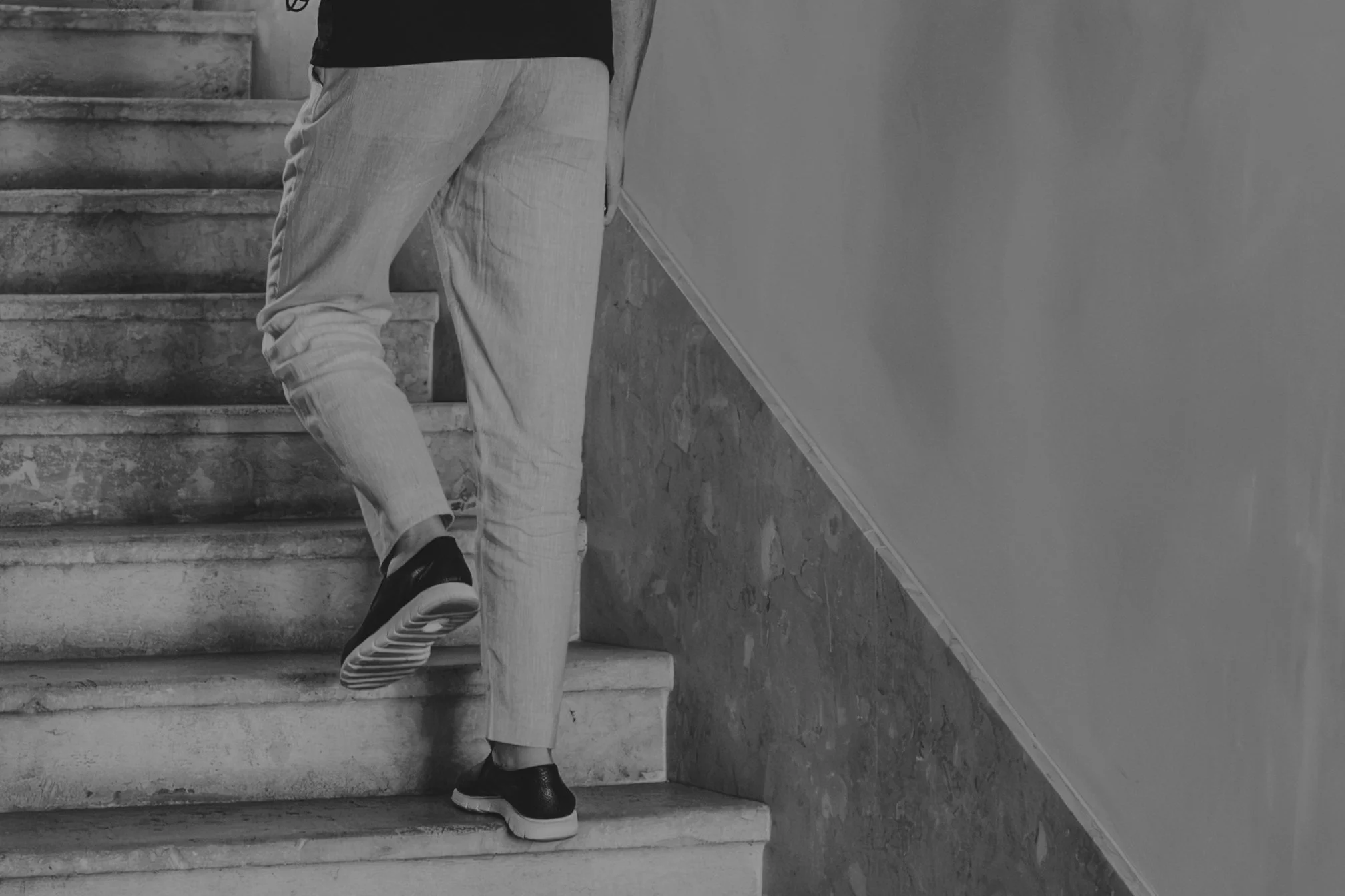 Person ascending marble stairs, wearing black shoes, light pants, and a dark shirt, with the photo taken from the side, focusing on the lower half of the body, against a plain wall.