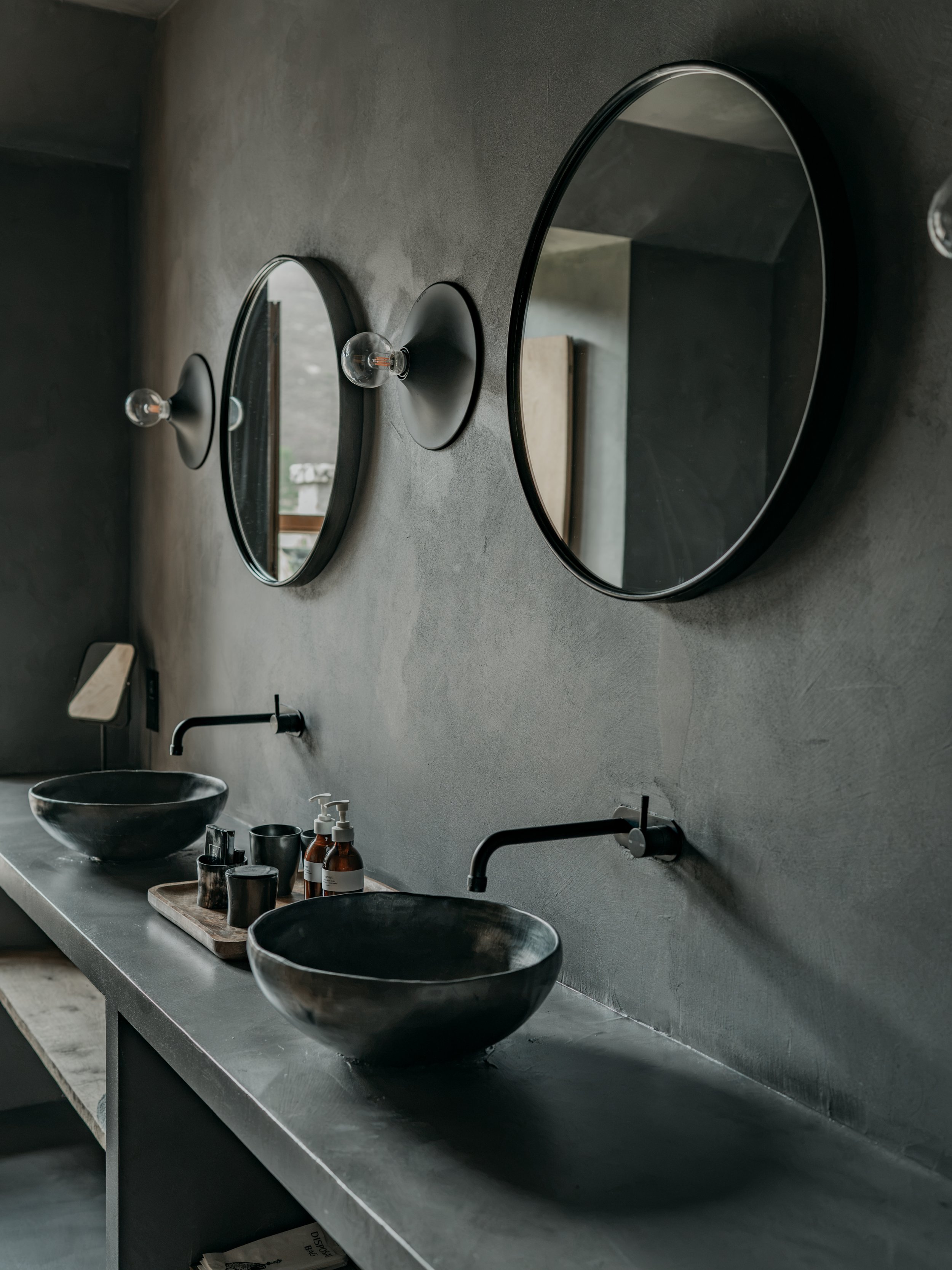 A modern bathroom with a concrete countertop, two black vessel sinks, and oval mirrors mounted on a textured gray wall. There are various toiletries and a wooden tray on the counter.