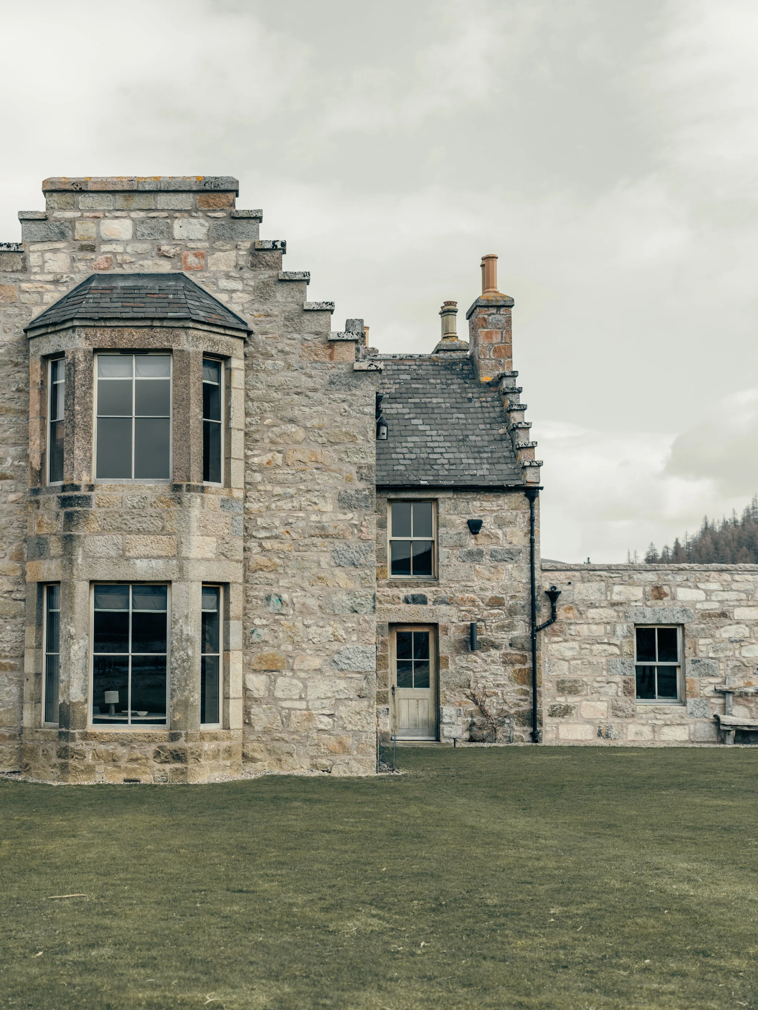 Stone house with large bay windows, multiple chimneys, and a manicured lawn under a cloudy sky.