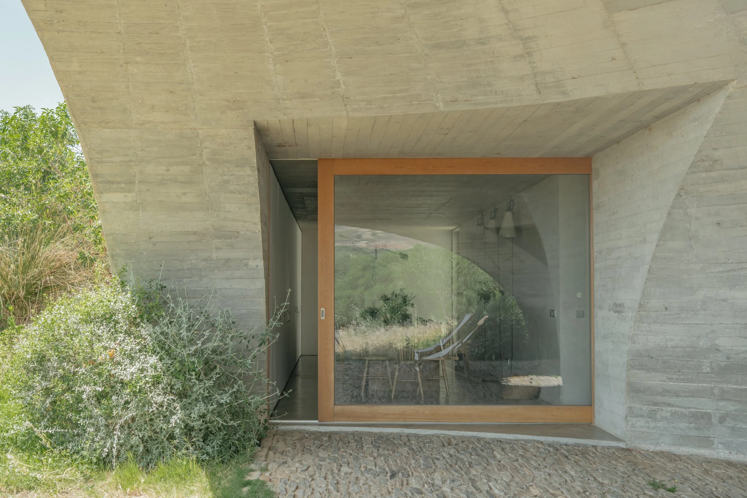 Modern concrete building with large glass sliding door, surrounded by greenery and trees.