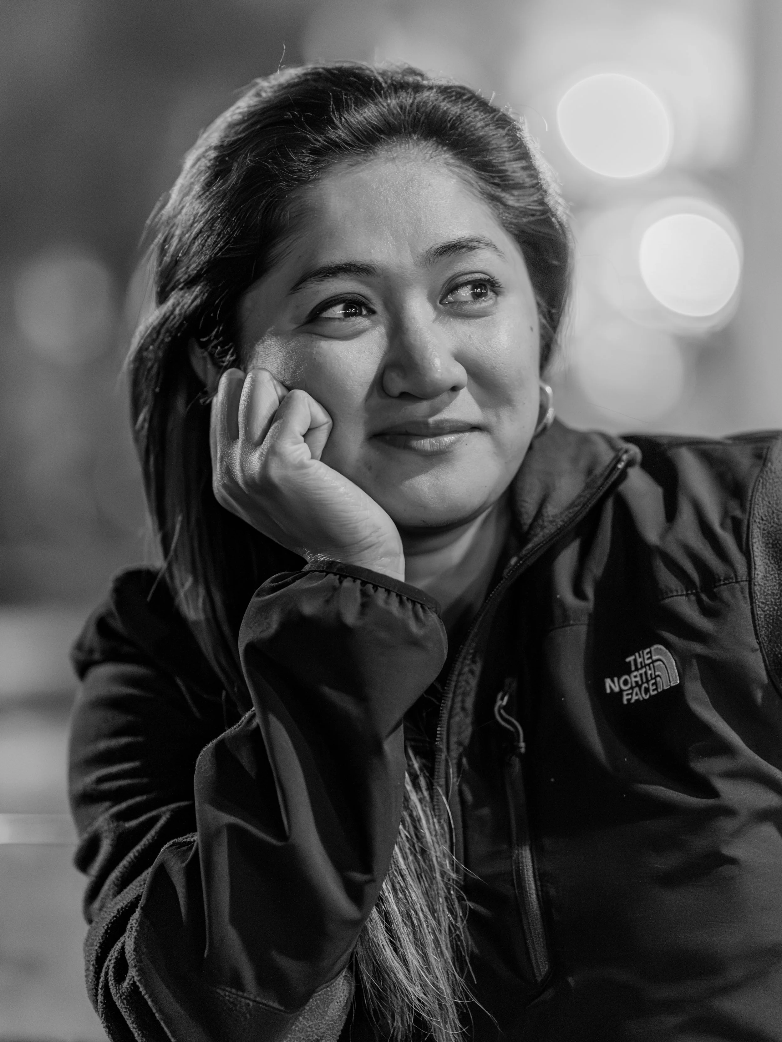 Black and white photo of a woman wearing a jacket, resting her head on her hand, with a blurred background of circular lights.