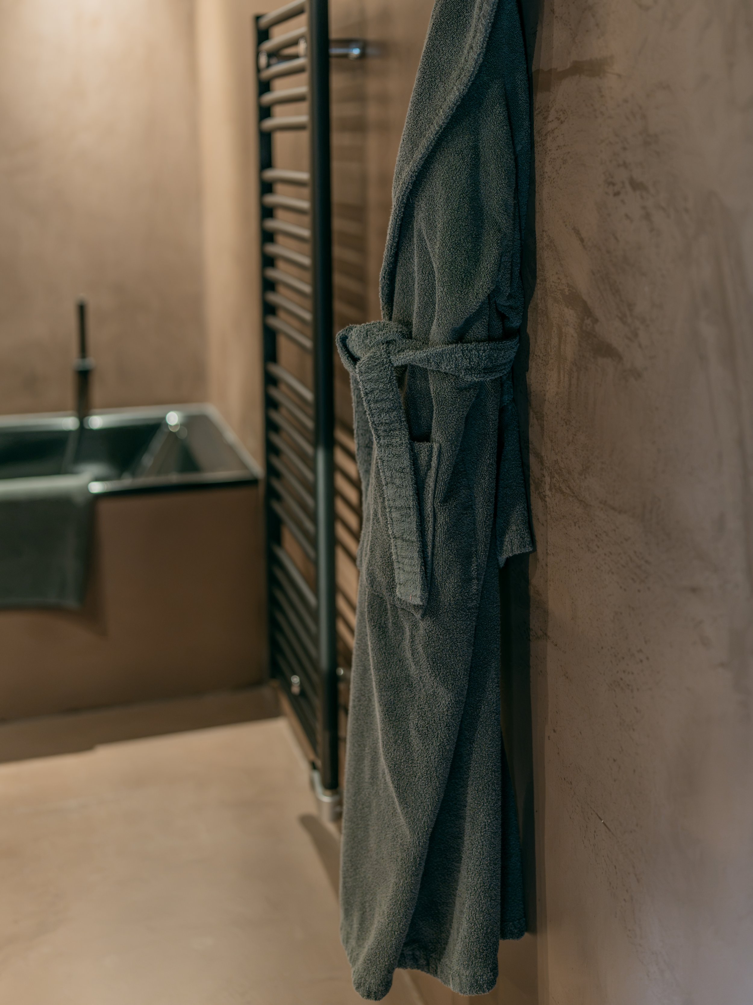 Gray bathrobe hanging on a hook in a bathroom with a towel rack in the background.