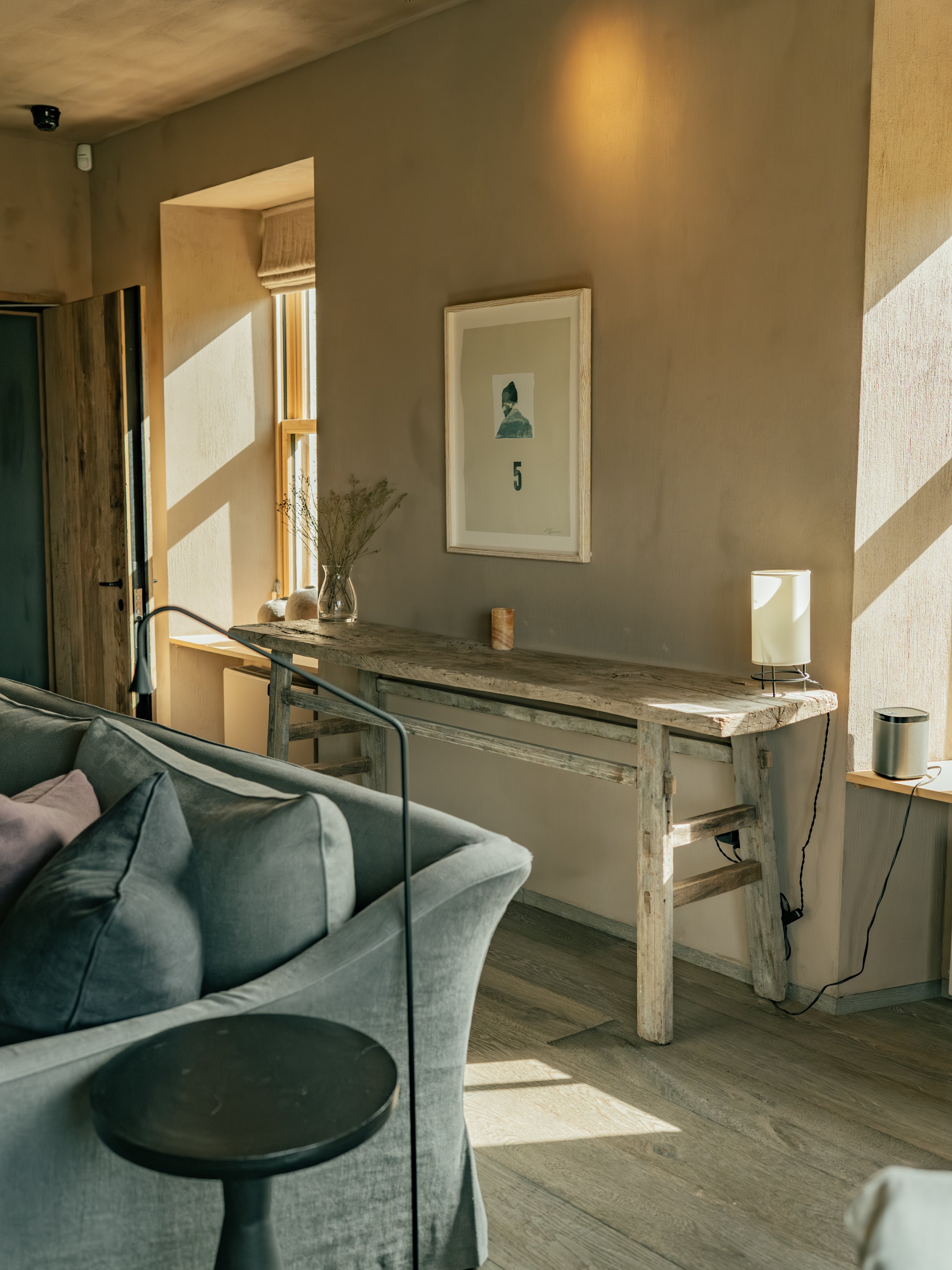Interior scene featuring a gray couch, a wooden console table with decor items including a vase with dried plants, a candle, and a lamp; a framed artwork with the number 5 on the wall; natural sunlight casting shadows; and a window with a beige Roman