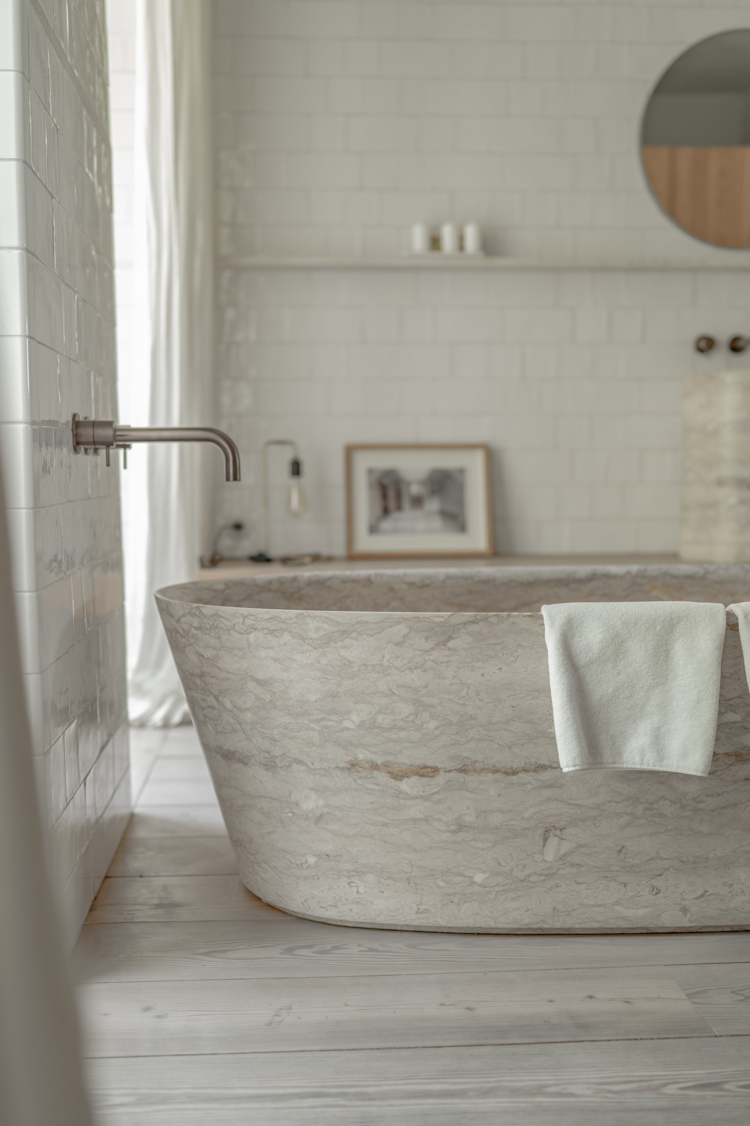 A white marble bathtub with a white towel hanging over its edge, situated in a minimalist bathroom with white brick walls, a round mirror, and a framed black-and-white photo in the background.