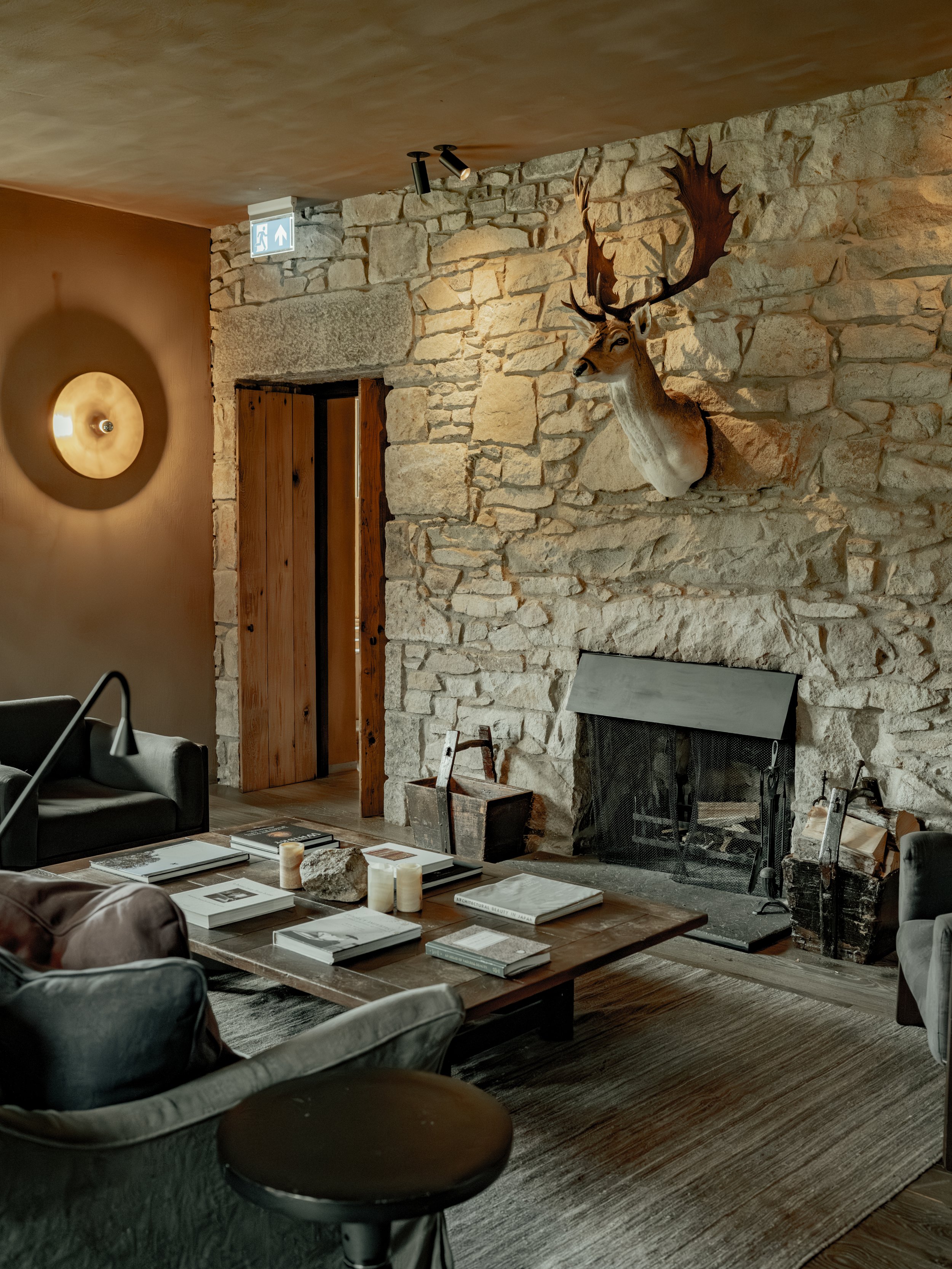 Interior of a cozy room with a stone fireplace, a mounted deer head with large antlers on the wall, a wooden door, a hanging lamp, a table with books and candles, chairs, and a rug.