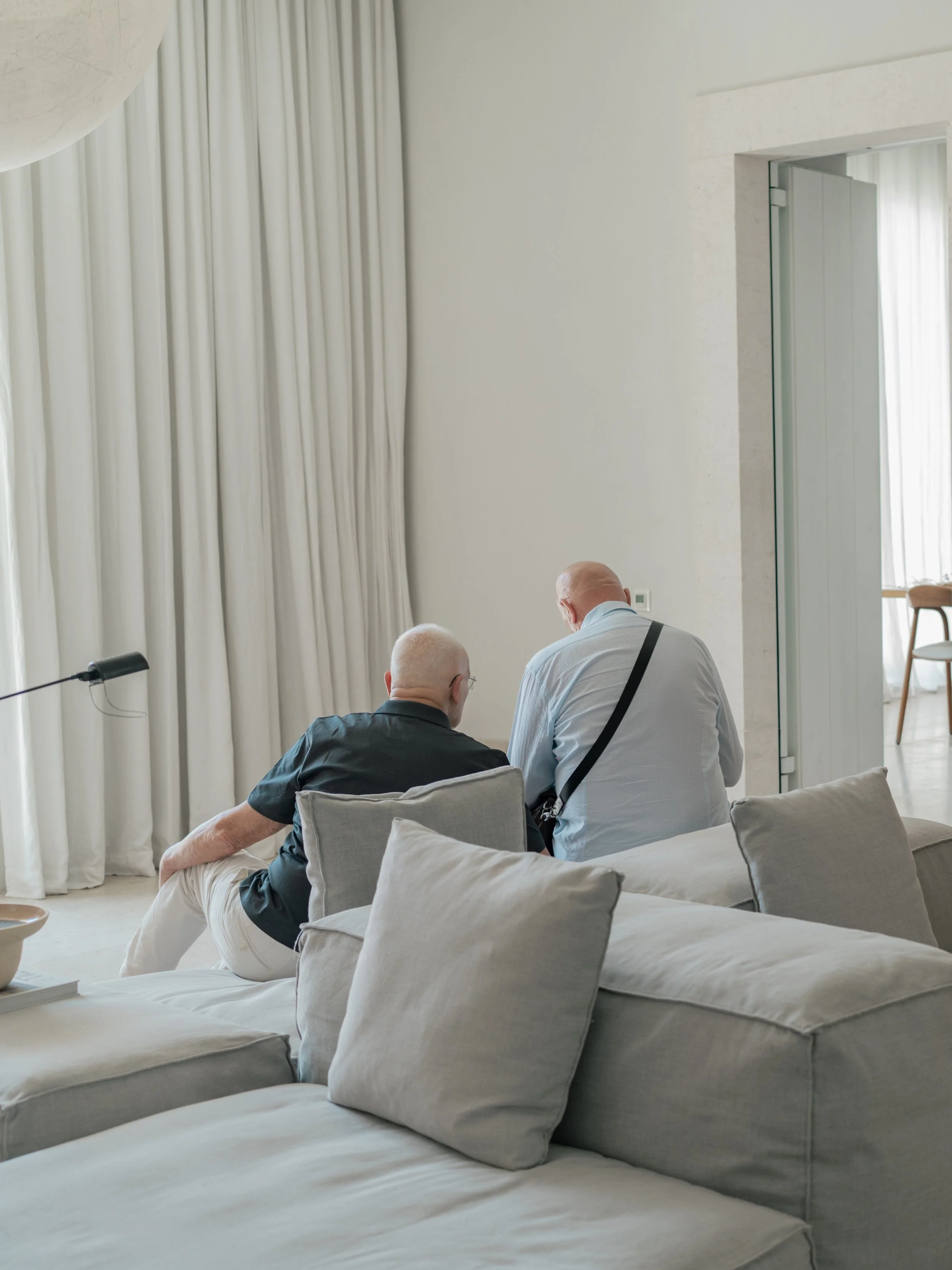 Two older men sitting on a sofa with beige cushions in a minimalist, well-lit living room with cream-colored walls, white curtains, and a doorway leading to another room.