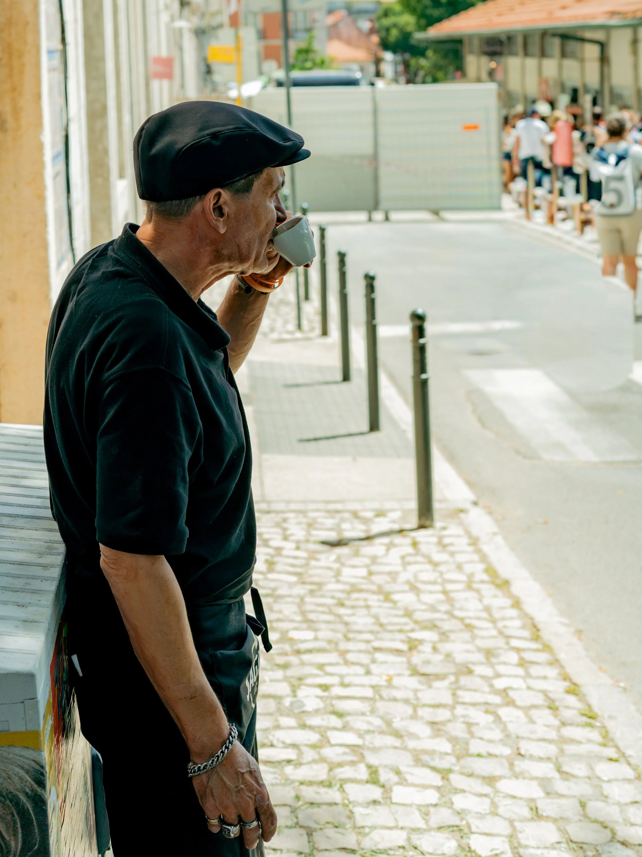 An elderly man with a black cap and black shirt standing on a sidewalk, drinking from a small white cup, with a blurred crowd of people and storefronts in the background on a sunny day.