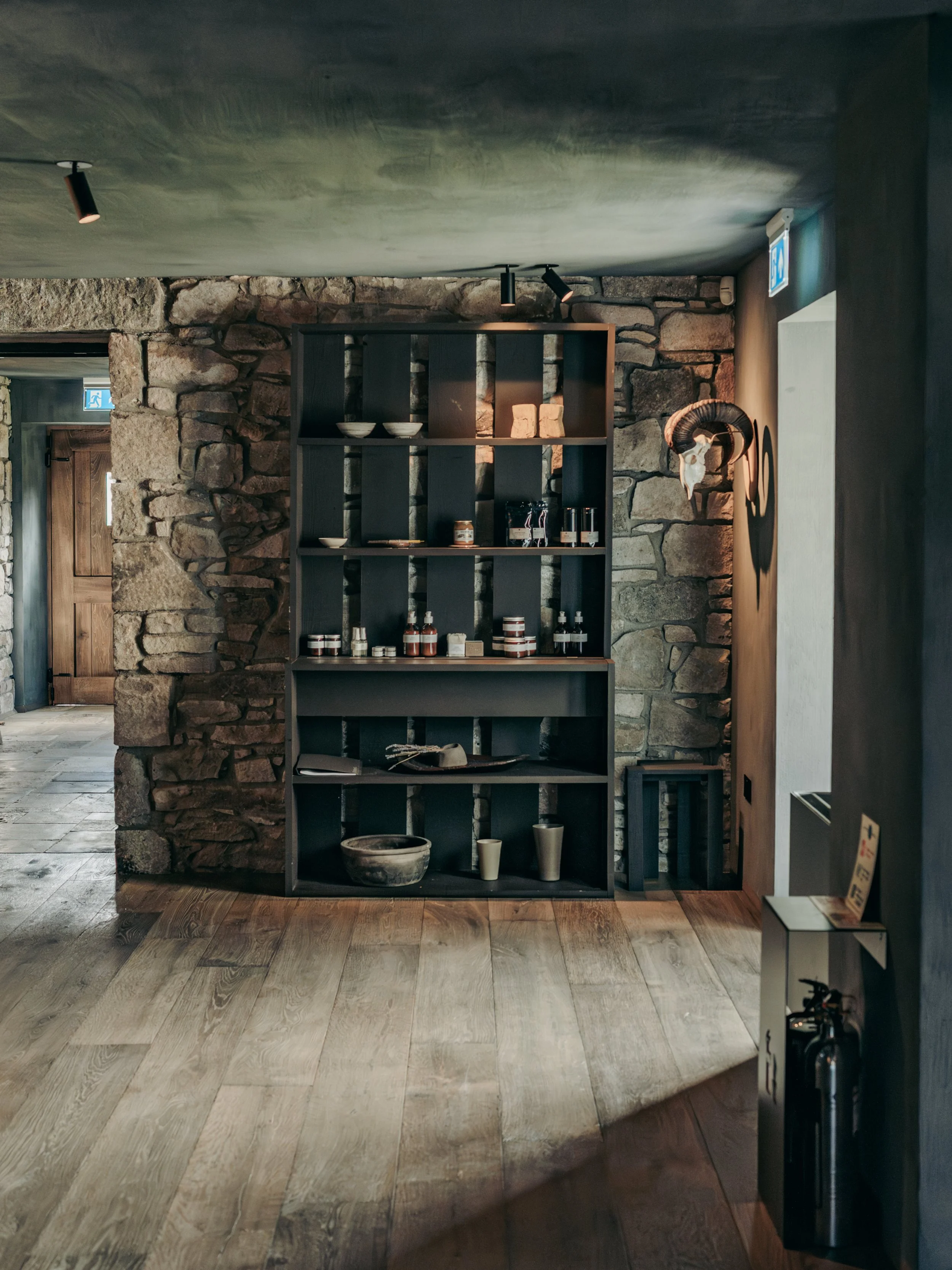 Interior of a rustic store or cafe with a stone wall, wooden flooring, a black shelving unit with bowls and jars, and a small mounted animal skull with horns.