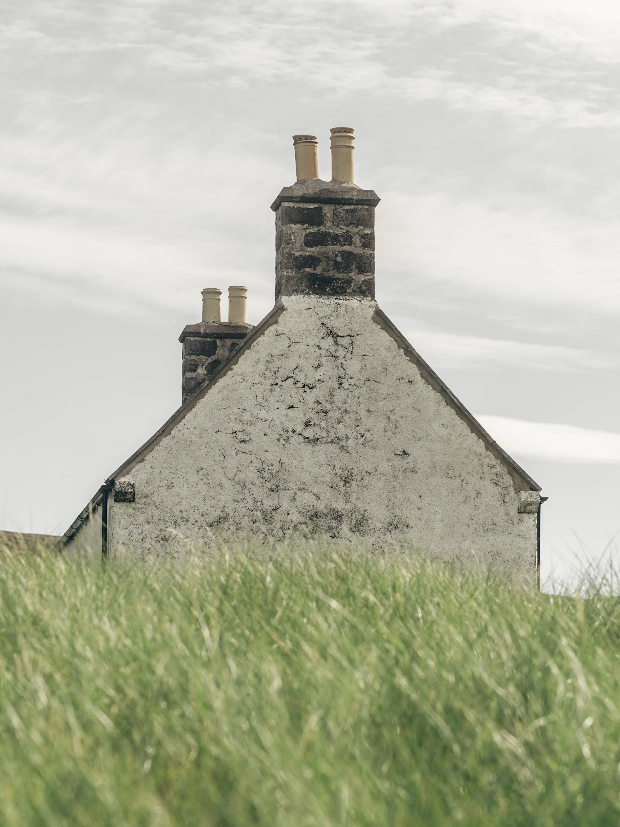 A house with a steep roof and two chimneys, viewed from ground level with grass in the foreground and a cloudy sky overhead.