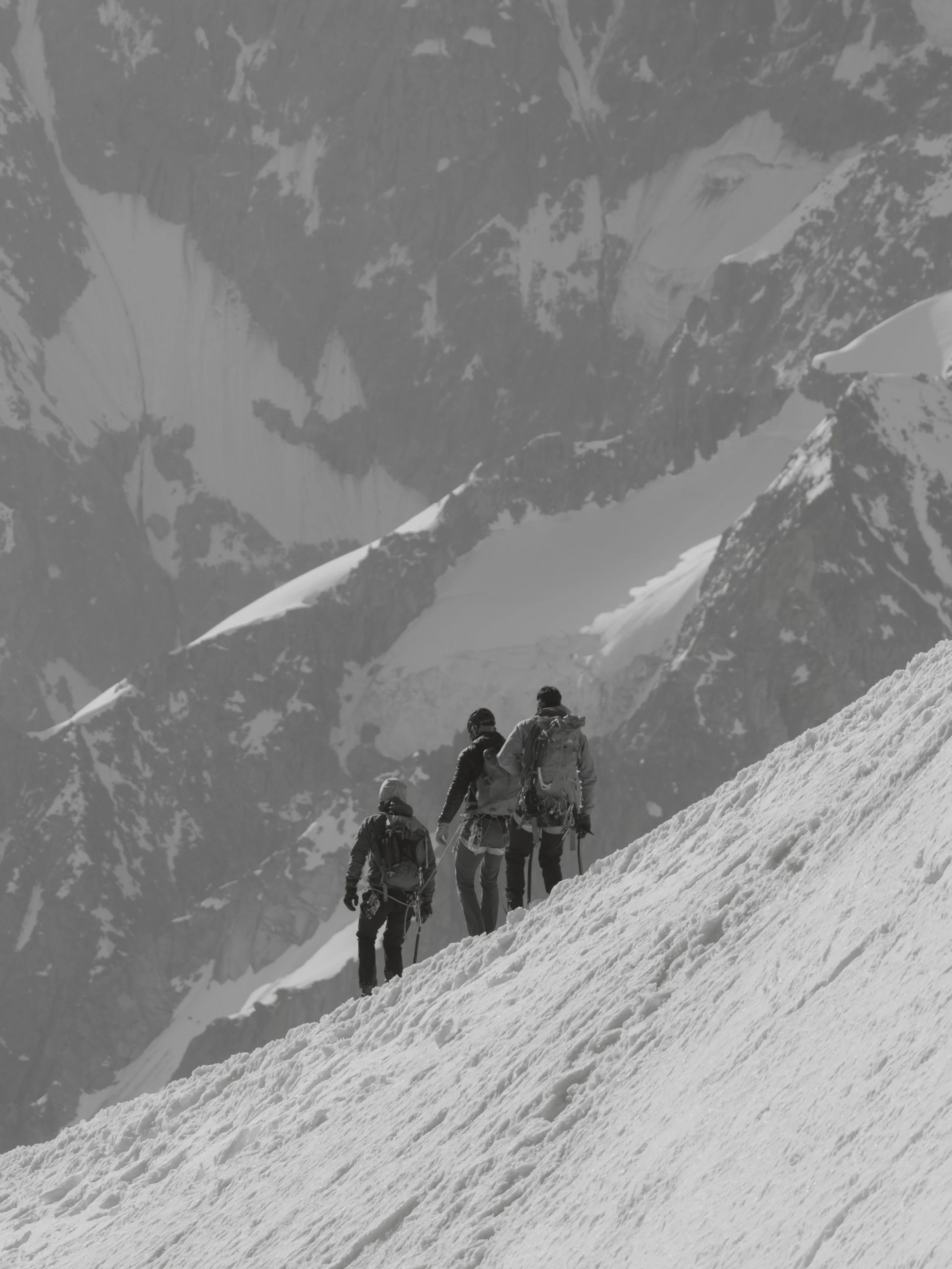 A group of six climbers, wearing winter gear and backpacks, ascending a snowy mountain slope with a backdrop of rugged snow-capped peaks.