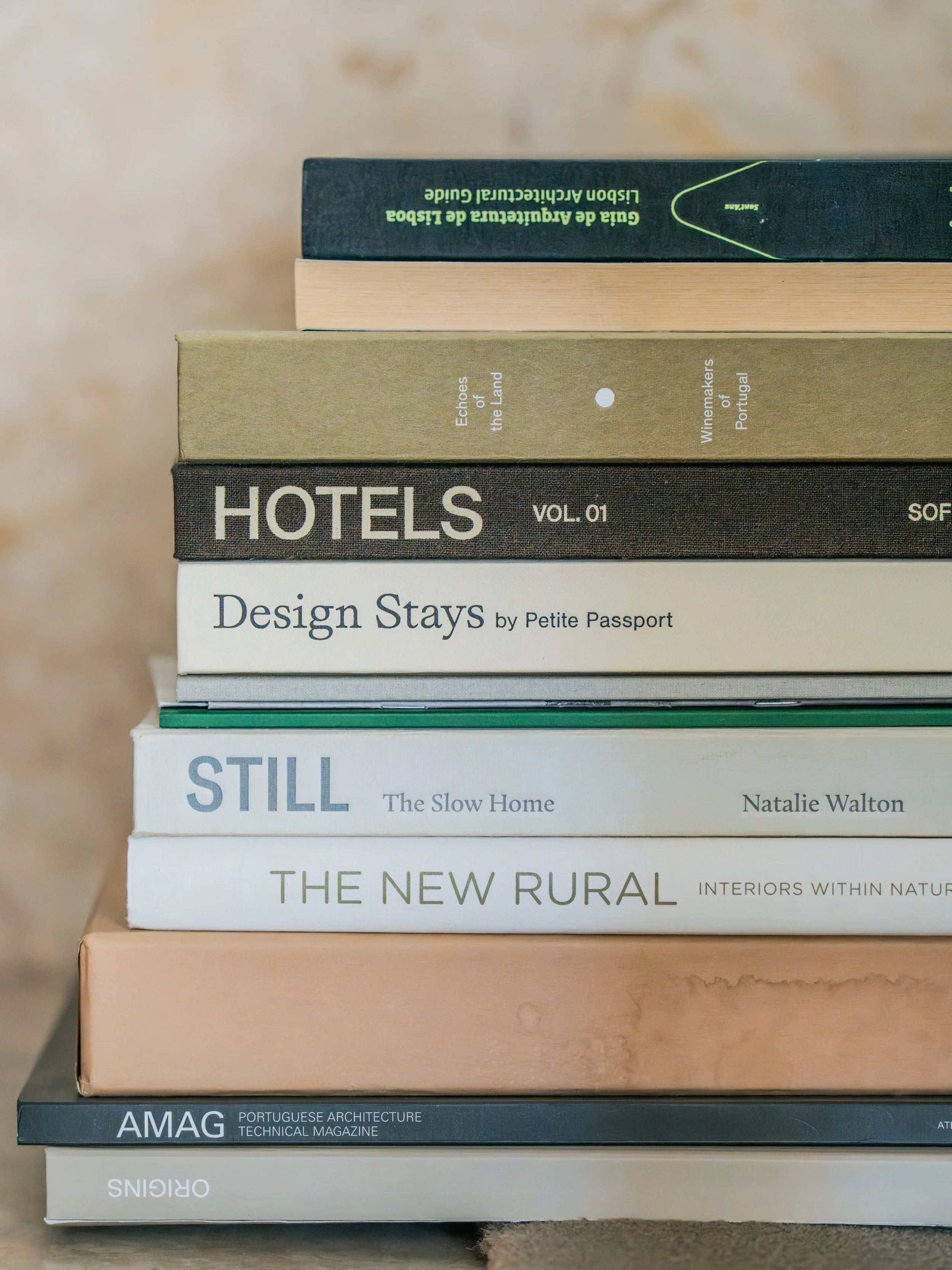 Stack of seven books on a surface, with titles including "The New Rural," "Still," "Design Stays," "Hotels," "Echres of the Land," and "Guia de Agricultura de Lisboa de Lisboa e Aricultural Guide."