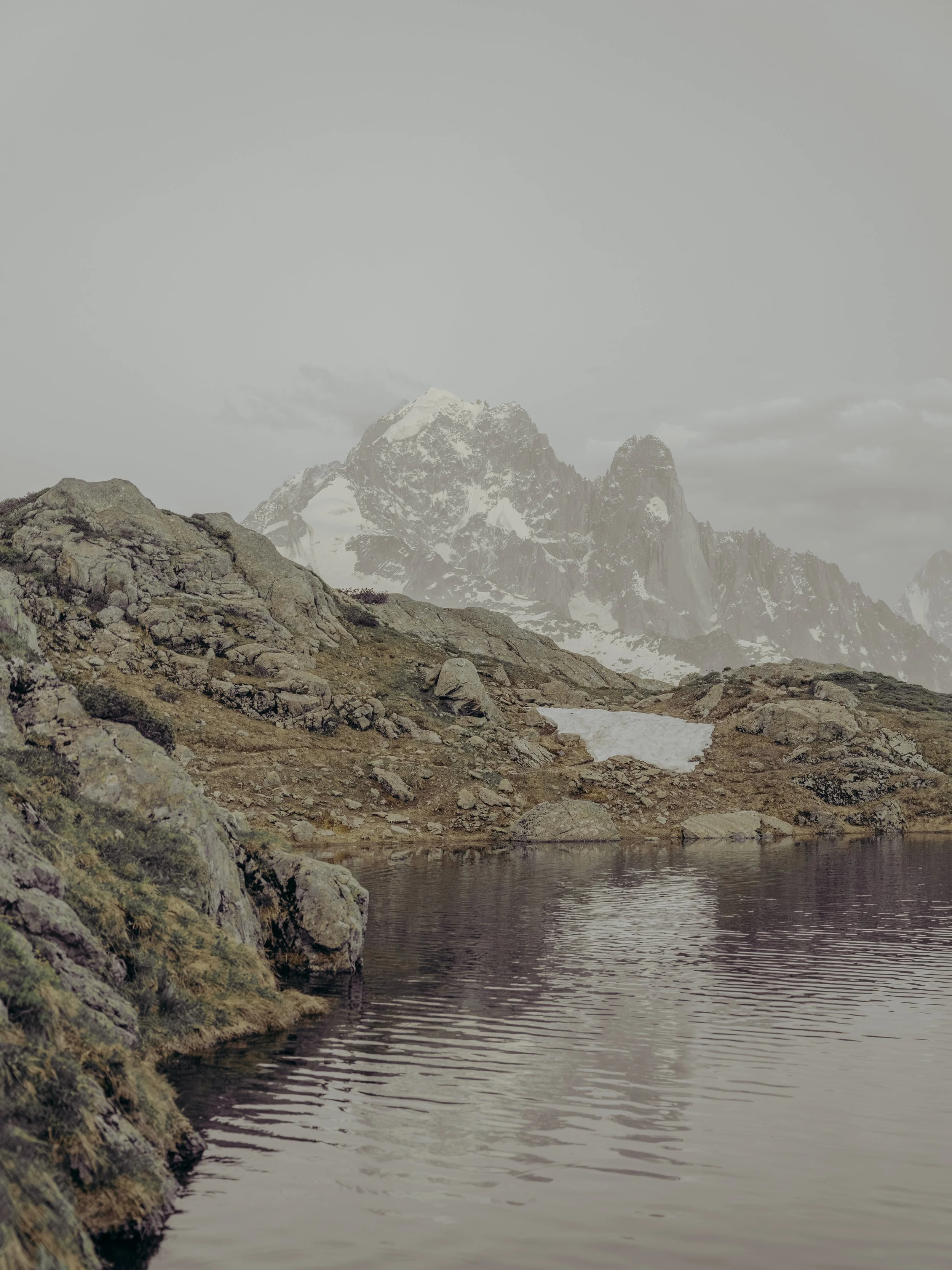 A mountain landscape with snowy peaks, rocky terrain, and a calm lake reflecting the scenery.