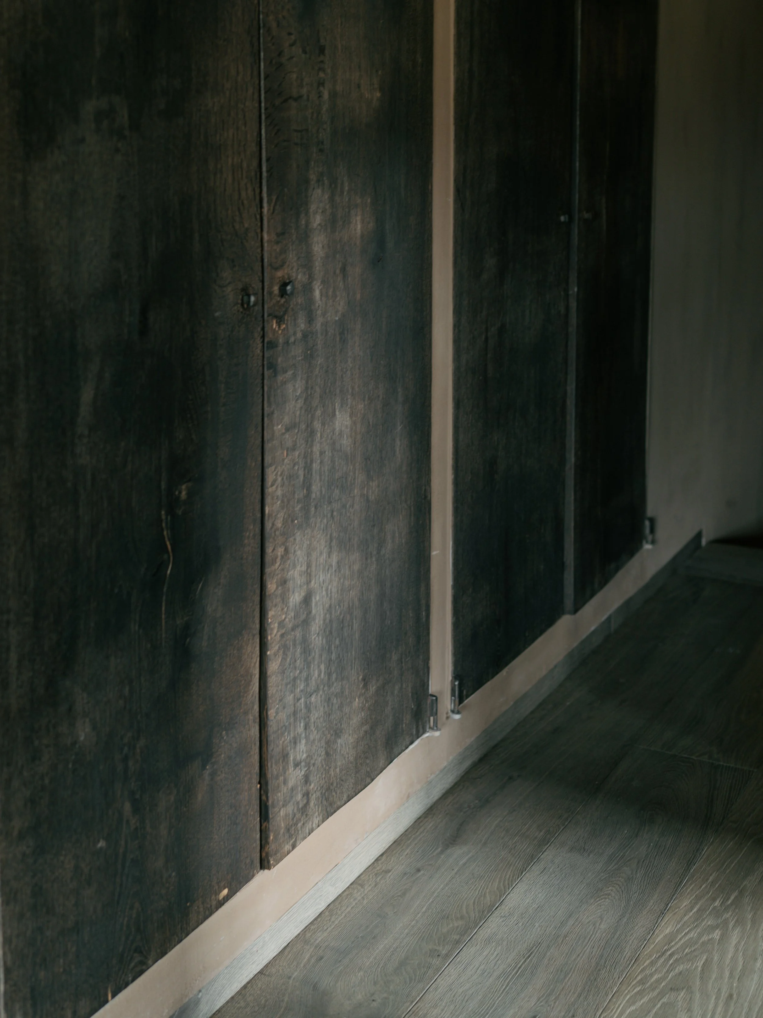 Black wooden panel wall with beige baseboard and dark wood flooring.