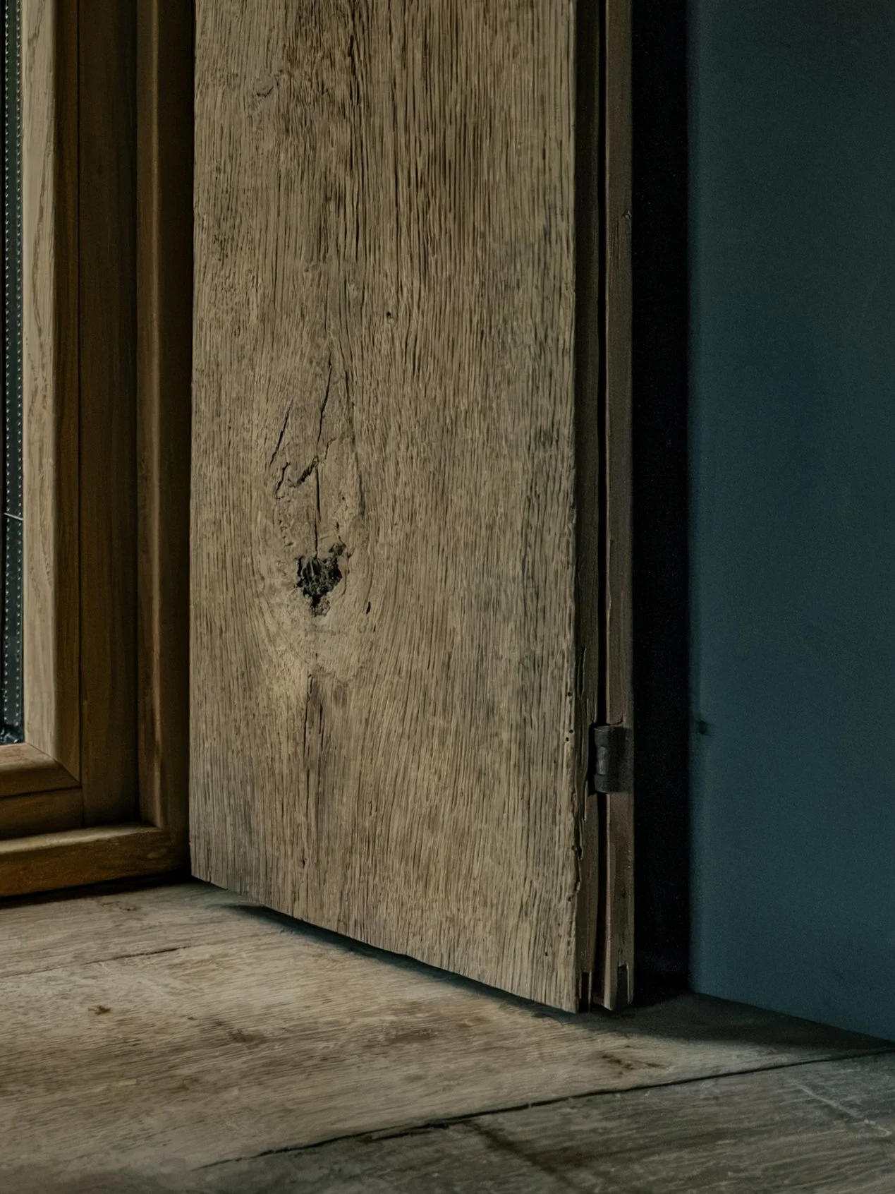 Close-up of a wooden door with visible grain, knots, and cracks, partially opened with a dark doorframe and wall to the right.