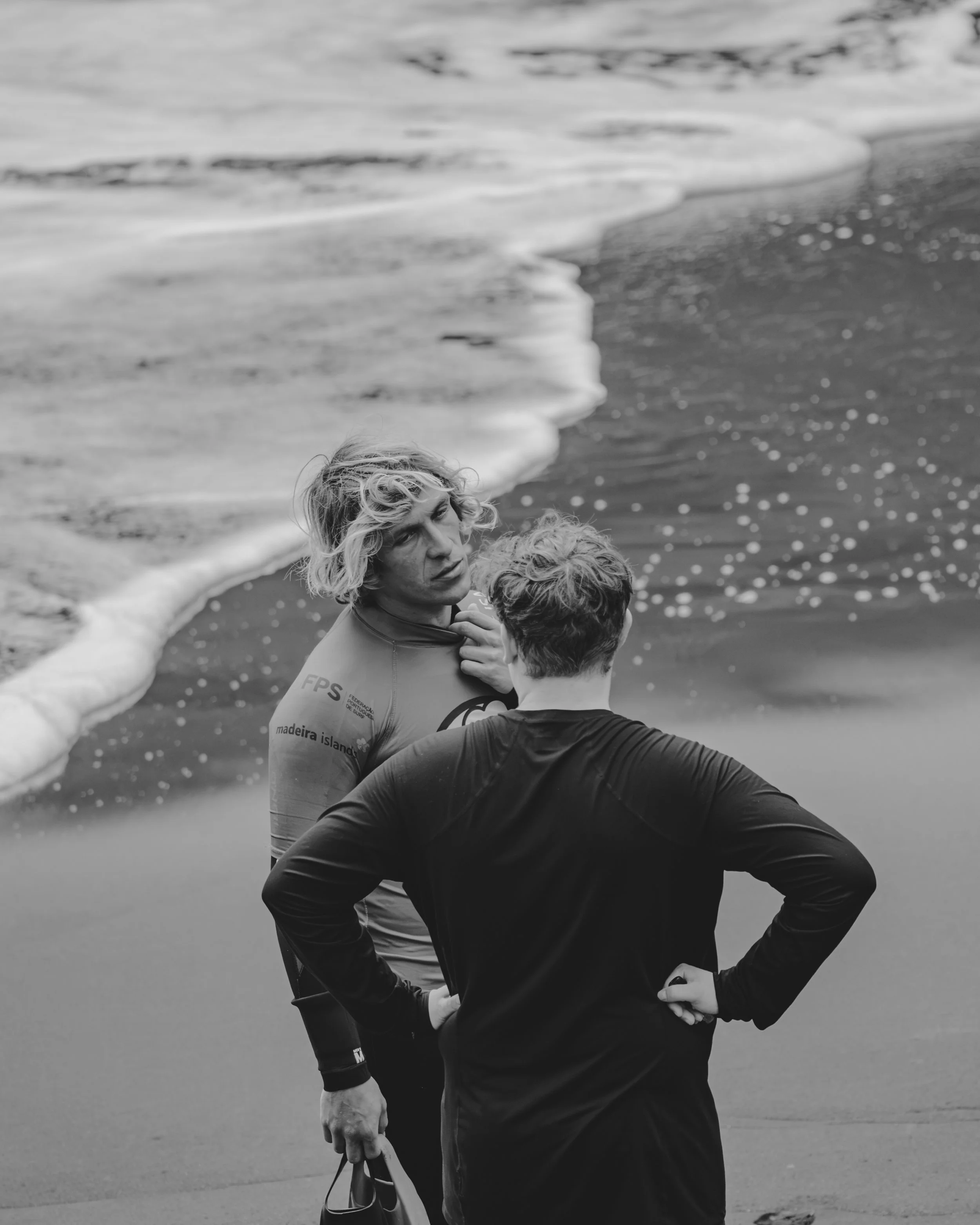 Two women on a beach, one with curly hair, the other with short hair, talking near the water's edge.