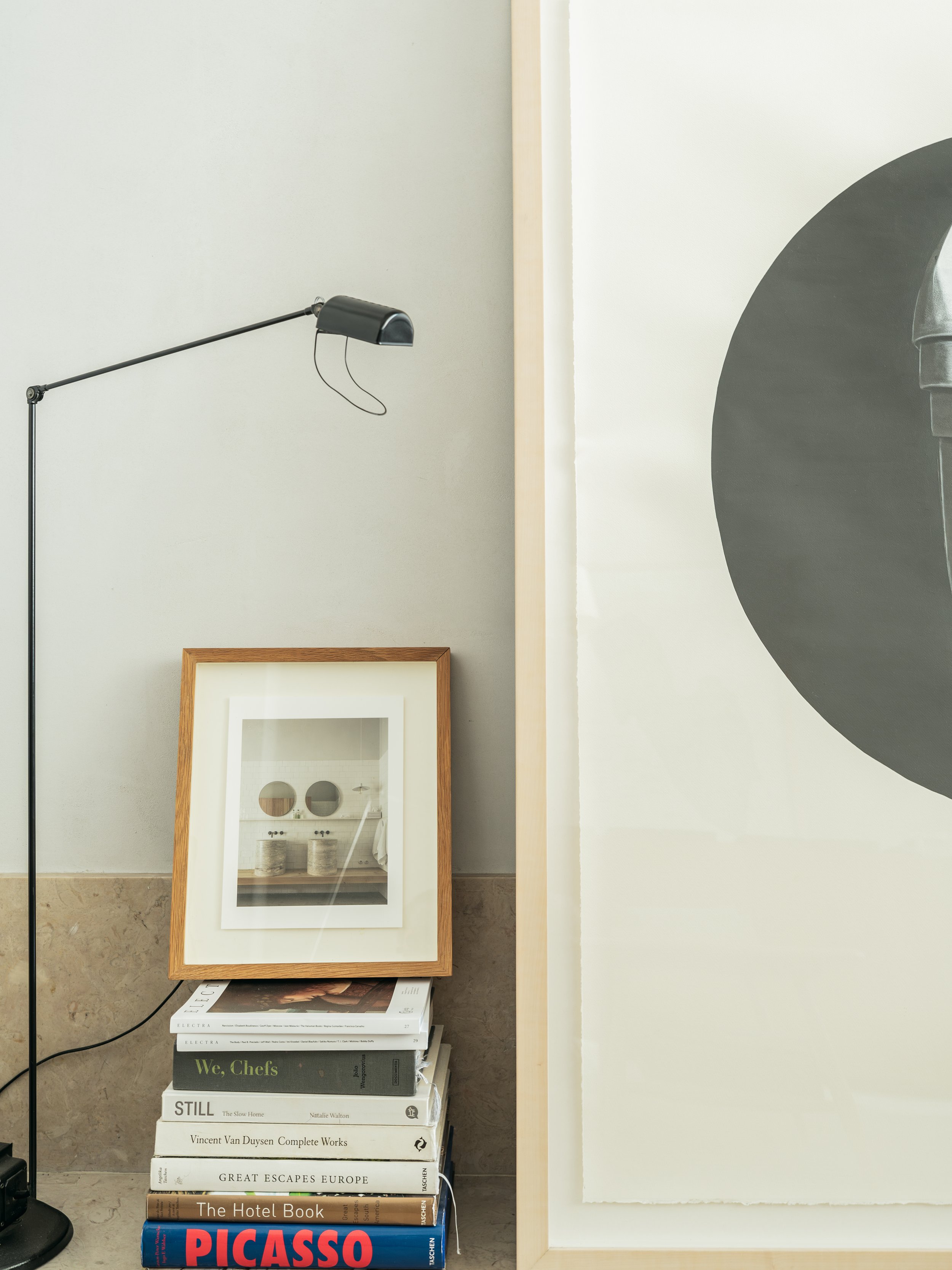 A stack of art, design, and travel books with a framed photograph of a minimalist interior scene, leaning against a wall, on a beige marble surface. A black desk lamp is nearby.