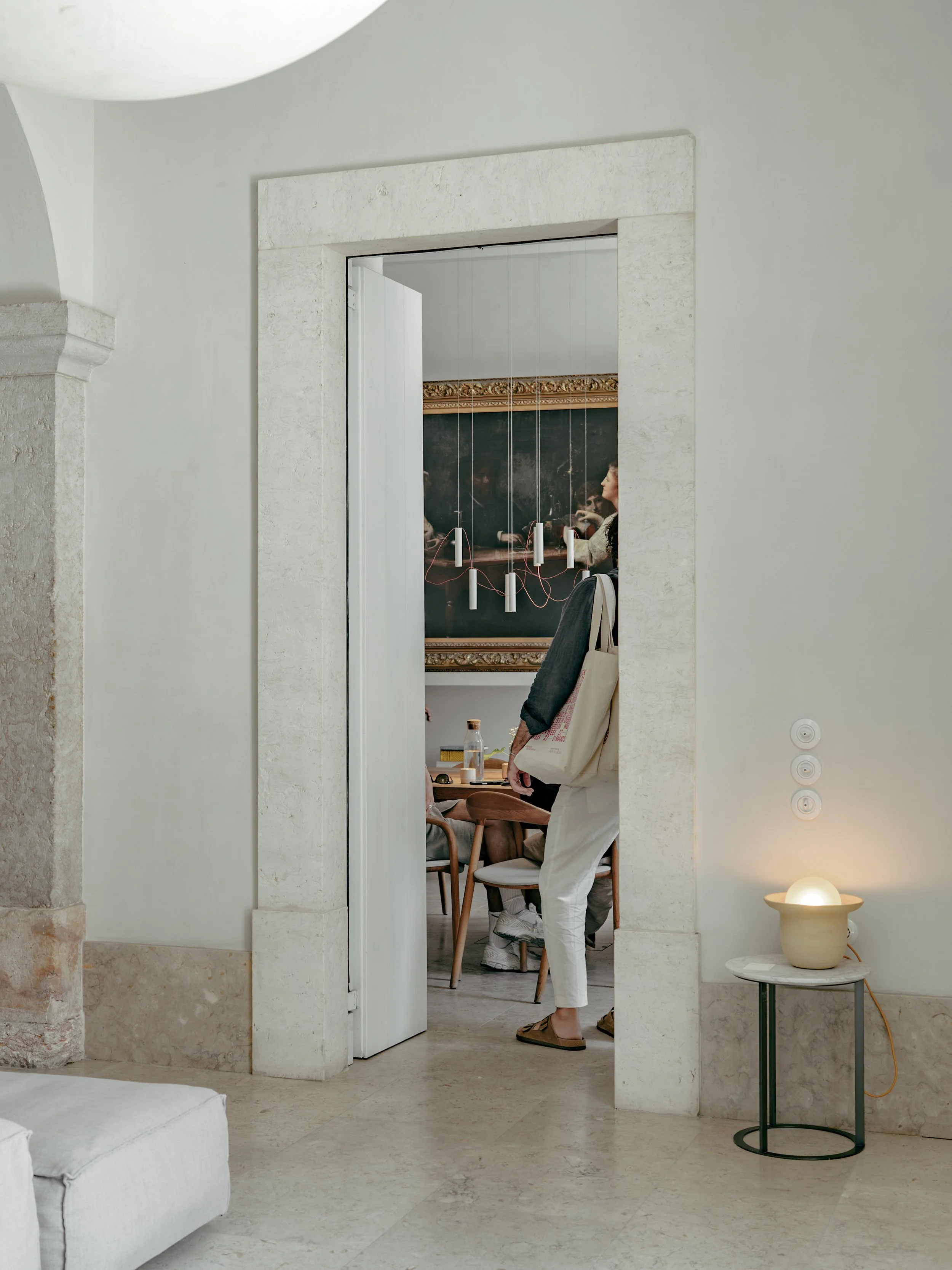 A person wearing white pants and slippers standing in a doorway, looking into a room with a blackboard and hanging pendant lights, while another person is seated at a table inside.