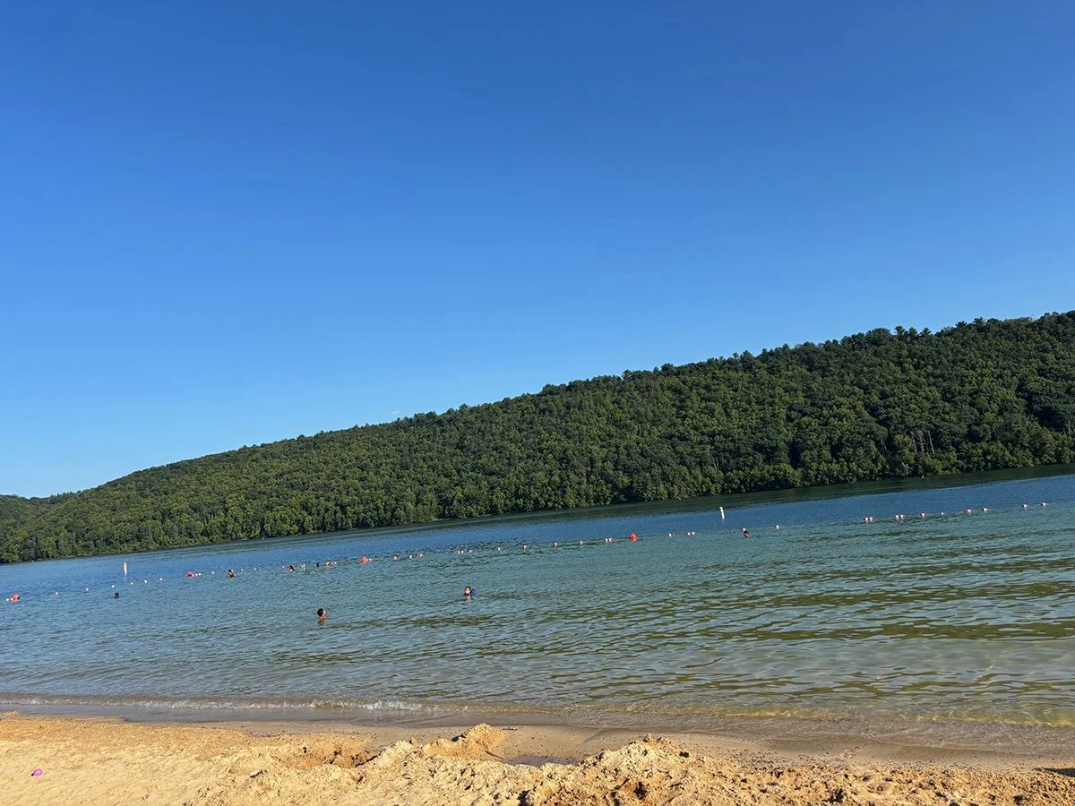 Scenic image of the swimming area at Beltzville Lake.  Clear blue sky, blue water with small waves, warm sand, and a mountain of green trees in the background.