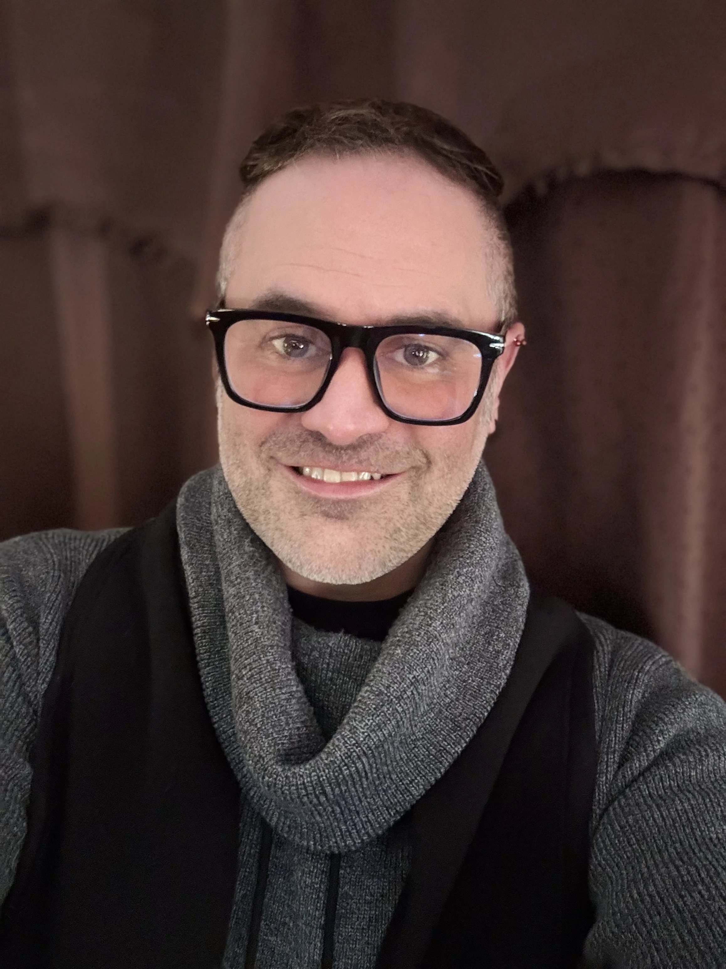 Headshot of the provider, Jonathan M. Blackwell-Rodgers Guest with a dark background.  He is looking dapper wearing black-rimmed glasses and a gray sweater.