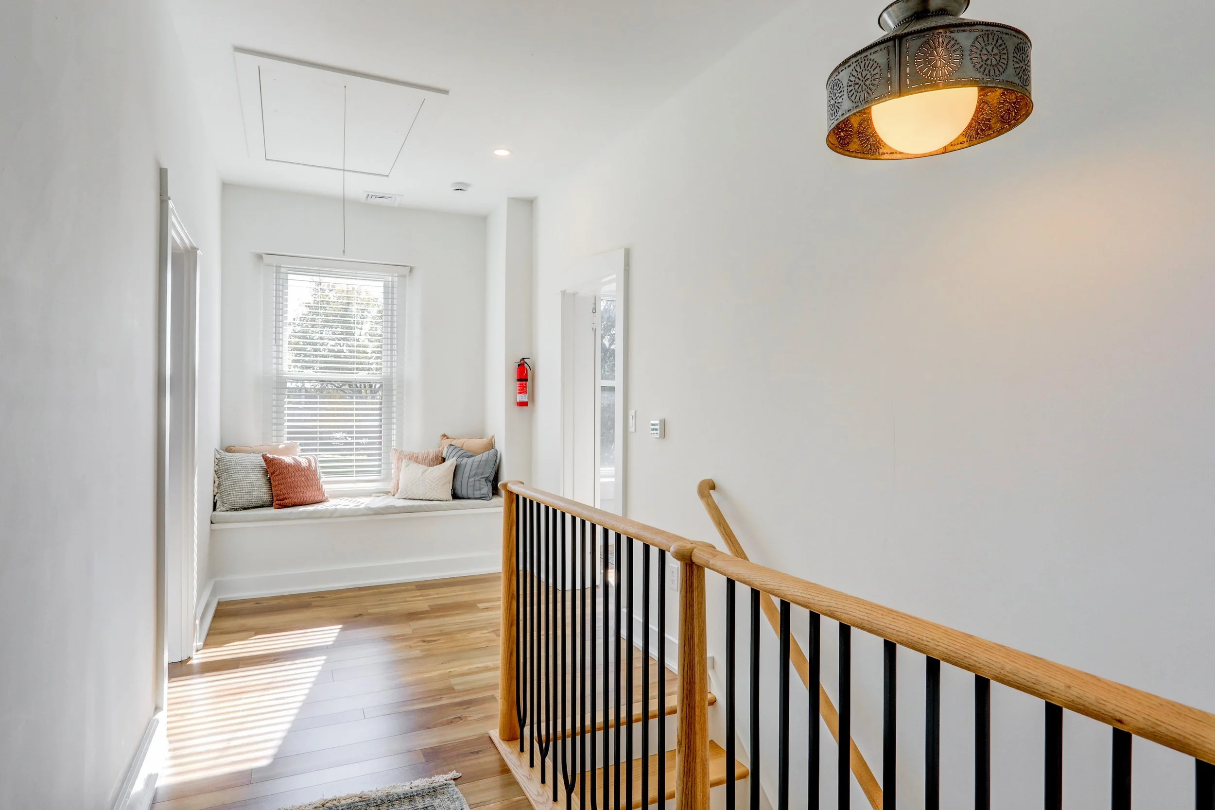 A bright, white residential space with a staircase and a window seat with pillows. Natural light filters through the window with blinds, and a fire extinguisher is mounted on the wall. The ceiling has an interior hatch, and an elegant ceiling light f