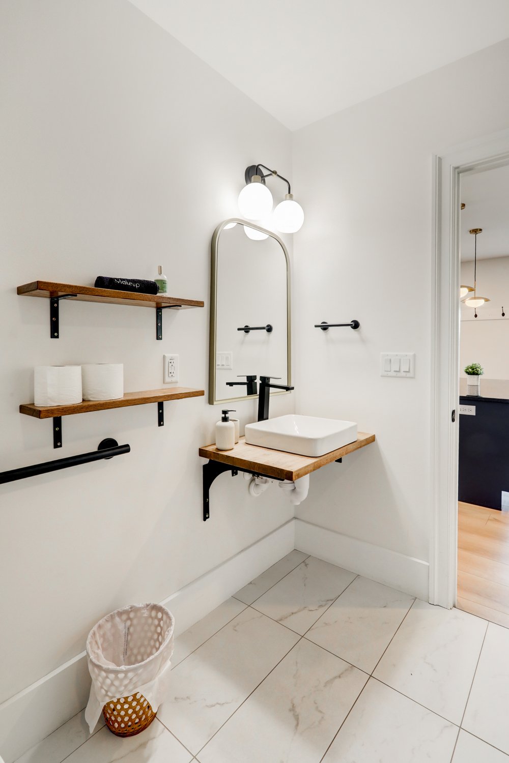 A modern minimalist bathroom with white walls, a rectangular vessel sink on a wooden countertop, a tall mirror, black fixtures, and wooden shelves with toiletries, illuminated by a three-bulb light fixture.