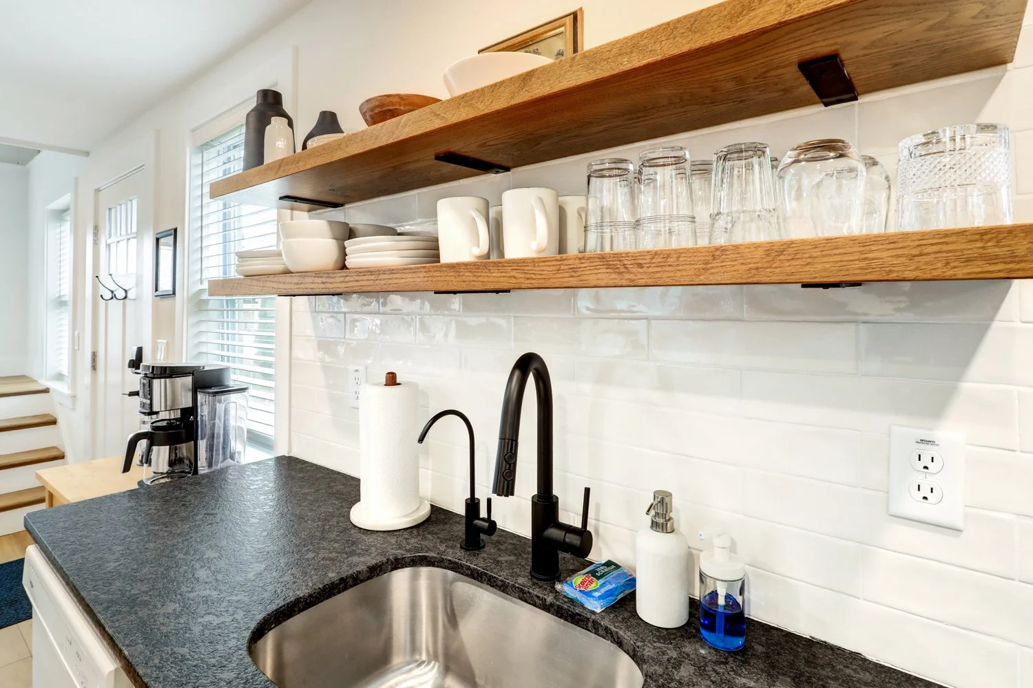 Kitchen with open wooden shelves holding dishes and glasses, black sink and faucet, paper towel roll, soap dispensers, coffee maker, and a window with blinds.