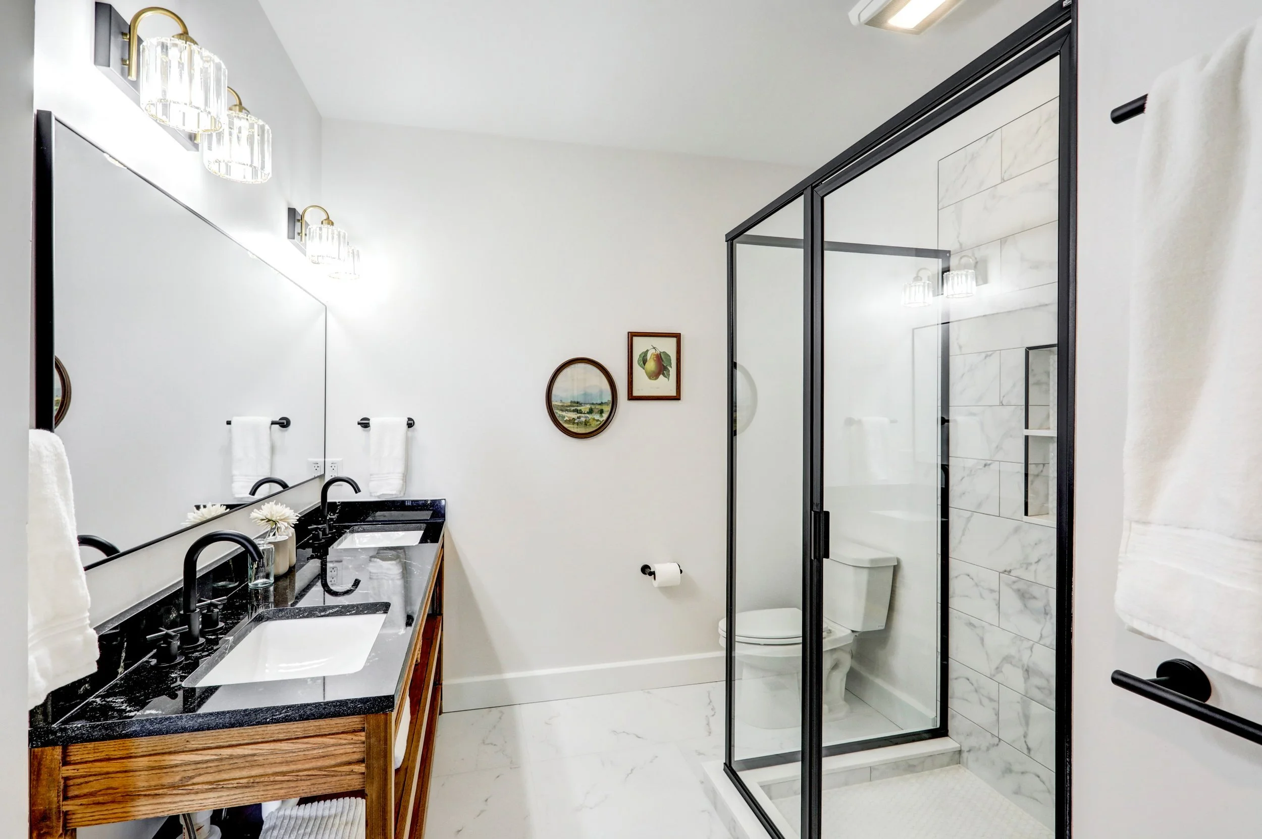 Modern bathroom featuring a double vanity with a black countertop and wooden base, wall-mounted mirrors, white towels, wall sconces, a framed pear print and landscape painting, a glass-enclosed shower with marble tiles, and a white toilet.