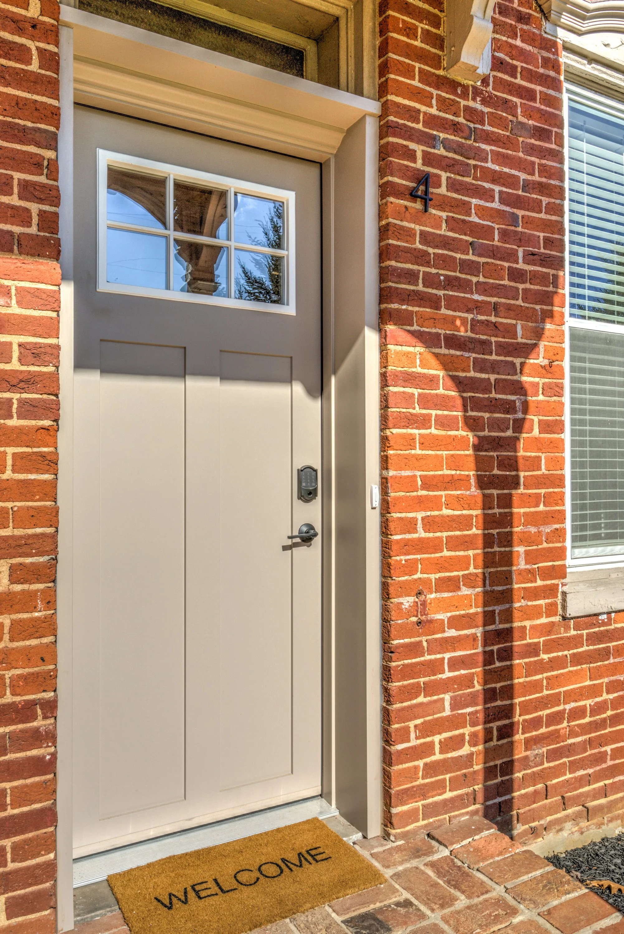 A front door with a large window on top, set in a brick house with a brick step and a welcome mat on the porch. The door has a digital lock, and a house number '4' is attached to the brick wall next to it. The door is framed with beige trim, and ther