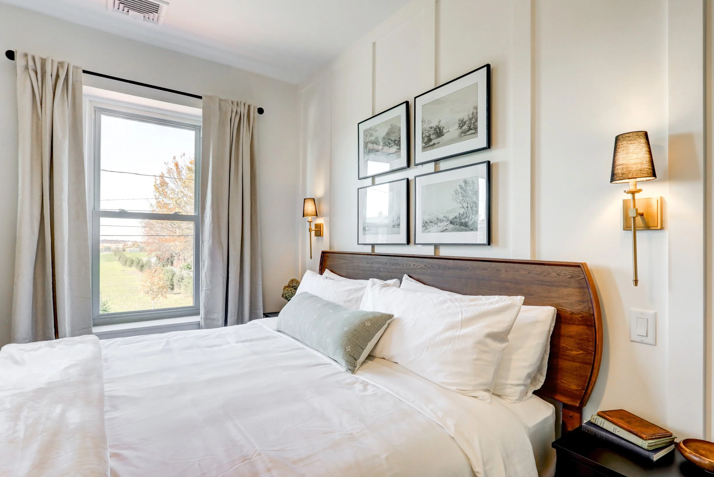 A cozy bedroom with white walls and a wooden headboard. There are four black-framed landscape artwork pieces hanging above the bed. Two gold wall sconces with black shades are mounted on either side of the artwork. A large window with white curtains 